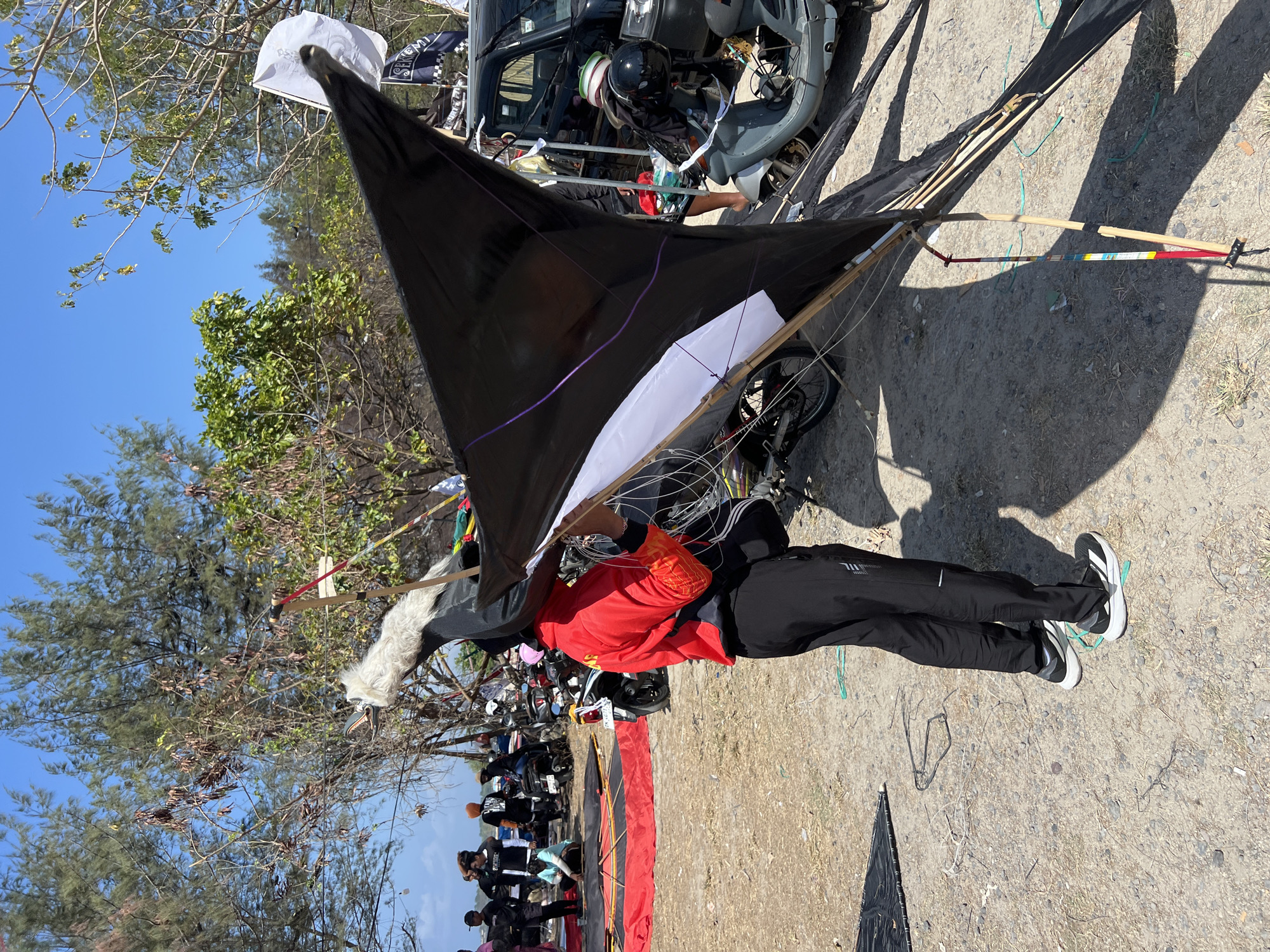 In Denpasar, Indonesia, on a sunlit beach with sparse vegetation, a person in a red jacket and black pants is assembling a large black and white kite. The kite has a distinctive white, fur-like adornment at its front, resembling a creature's head, and purple trim. In the background, several other individuals are visible, some near motorcycles and a black car. Red tarps are spread on the ground, likely for preparing kites or gathering materials. The ground is a mix of sand and gravel. Several motorcycles are parked nearby, suggesting transportation for the attendees. The overall scene depicts an outdoor activity or event related to kite flying.