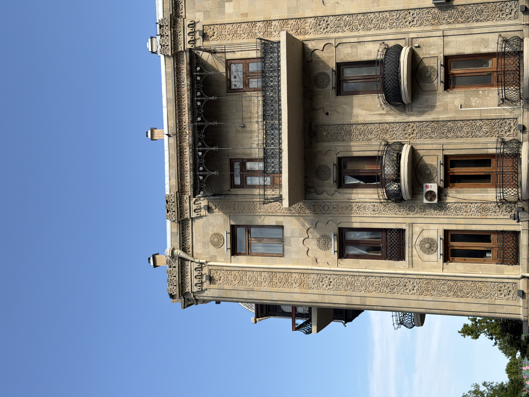 The image shows an ornate stone building with intricate carvings and multiple balconies under a clear blue sky. The architecture suggests a historical building, possibly in a city with such elaborate designs. The building is a multi-story structure, with wooden framed windows and doors. Several balconies with black wrought-iron railings are visible. The building's facade is decorated with geometric patterns and floral motifs. In the lower left corner, the tops of green trees are visible, indicating an outdoor setting. The lighting suggests it is daytime, with shadows cast on the building.