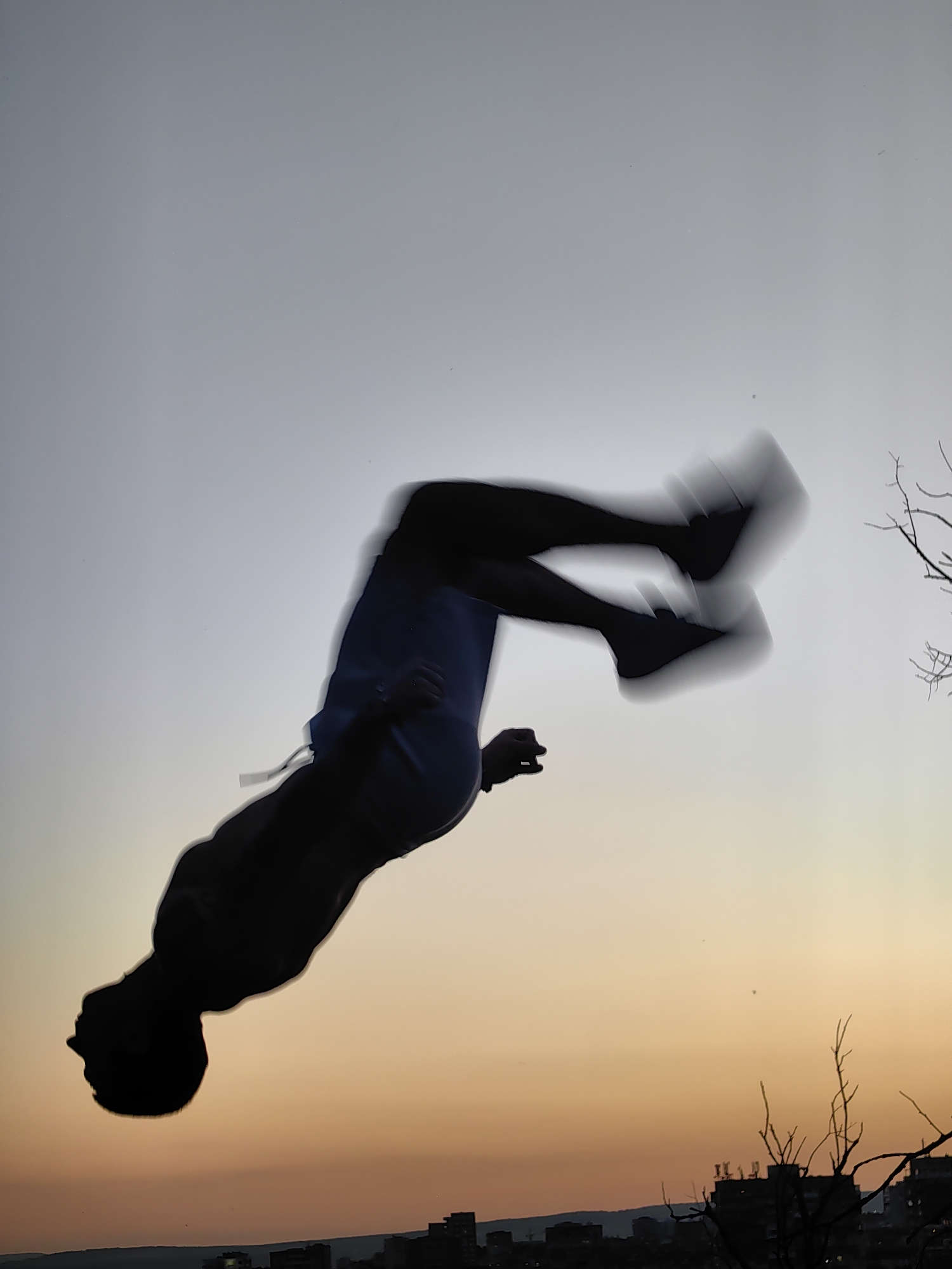 A silhouette of a person mid-flip against a gradient sky of pale gray to orange is captured. The person appears to be in motion, with limbs blurred. The background shows a hazy cityscape with buildings silhouetted against the horizon. Bare branches of a tree are visible in the upper right portion of the frame. The overall scene suggests an outdoor activity taking place at dusk. The location is Varna, Bulgaria.