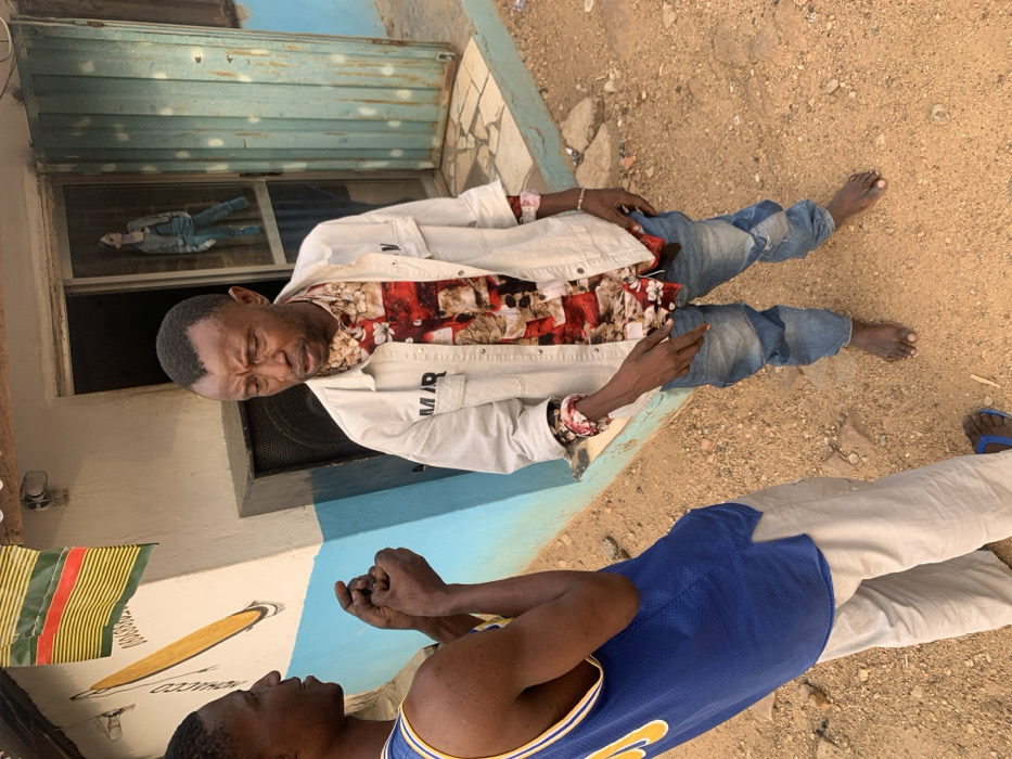 This image depicts two adult males interacting outdoors in Okene, Nigeria, against the backdrop of a building.
The man on the left, facing right, wears a blue basketball jersey with yellow accents and light-colored pants, completed by blue flip-flops. His hands are clasped at chest height, and his mouth is slightly open as if speaking or mid-sentence.
The man on the right, facing left, has a distinct V-shaped hairline. He wears an unbuttoned light beige over-shirt with "MIR" visible on the left chest, layered over a multi-colored, long-sleeved collared shirt with red, white, brown, and black patterns. He wears distressed blue jeans and is barefoot. A light-colored bracelet adorns his right wrist, and a small, dark object (possibly a phone) is held in his left hand. His facial expression is somewhat pensive, with furrowed brows, and he appears to be looking downwards and to his left, seemingly listening or reflecting.
The men stand on a dirt and gravel surface. Behind them, a building wall features a light-colored base with a horizontal blue stripe. A mural on the wall depicts a long, yellow, club-like object with black outlines, accompanied by partial text "COC..." and numbers "080808..." below it. Above the mural, a flag with horizontal red, yellow, and green stripes is visible. To the right of the mural, a dark rectangular speaker or utility box is situated next to a doorway. This doorway is fitted with a screen door, behind which a cutout or poster of a person in casual attire is visible. Further right, a light blue rolling metal door or shutter is partially open. The scene is illuminated by natural daylight.