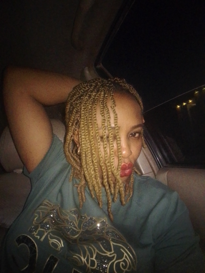 A dimly lit, close-up photograph captures a female individual seated inside a vehicle at night. The subject, appearing to be in a passenger seat, has light-brown skin and wears a dark teal or green short-sleeved top featuring a prominent, glittery gold and black patterned design on the front. Her hair is styled in blonde kinky twist braids, with several strands falling forward, partially obscuring the left side of her face. Her visible right eye is directed towards the camera, and her lips are painted red. A small, gold-colored hoop earring is present on her right ear. Her right arm is raised, with the hand positioned behind her head, resting against the light-colored car upholstery. The car's interior is visible, including a section of the headliner and a window frame on the right, through which distant, faint lights are discernible against the dark exterior. The depicted scene is described as taking place in Barnawa, Nigeria.