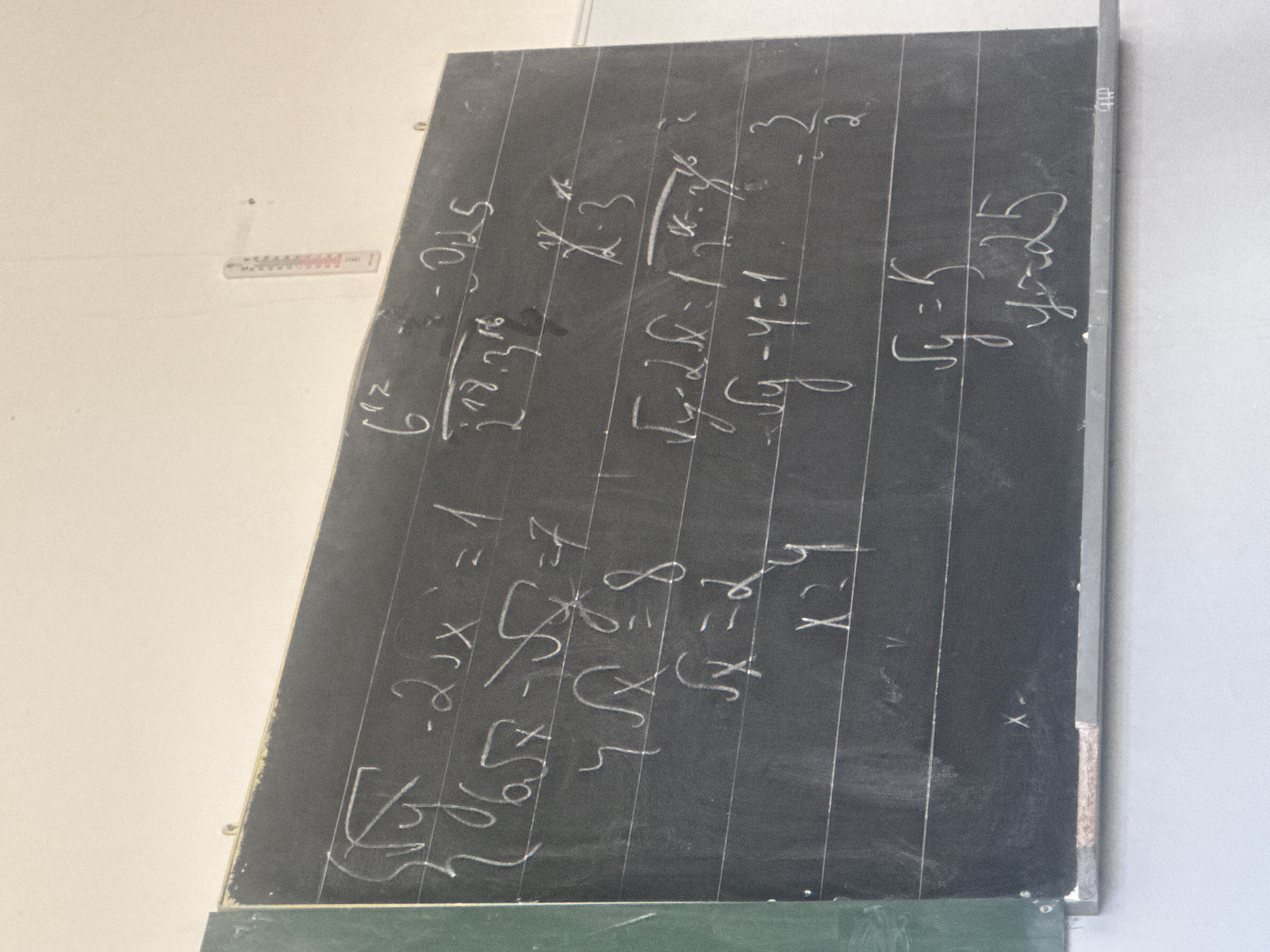 The image displays a dark gray chalkboard covered with mathematical equations and expressions written in white chalk, suggesting an educational setting. The chalkboard is angled, appearing to lean upwards and to the right, and is mounted against a plain, light-colored wall. A small, white object with red markings, possibly a thermometer or ruler, is partially visible on the wall to the left of the board. A strip of green surface, likely the bottom edge of the chalkboard or a chalk tray, is visible at the very bottom of the frame.
No people are visible in the image. The chalk writing indicates a session of problem-solving or a lesson. The equations involve algebraic terms, including exponents, square roots, and variables 'x' and 'y'. Some clearly decipherable expressions include: "- 0.25", "6^-2", "2^18 * 3^16", and "- 2x = 1". There are also several instances of root equations, such as "√y - 4 = 1", which is followed by "√y = 5" and subsequently "y = 25" written multiple times. Other equations include "√x = 8" and "x = 9", suggesting either different problems being solved simultaneously or steps with corrections. The writing style is somewhat casual, typical of chalkboard notes, with some lines appearing partially erased or overwritten. The scene depicts a classroom or study environment, likely indoors in Yerevan, Armenia, though no specific local cues are visible beyond the written content.