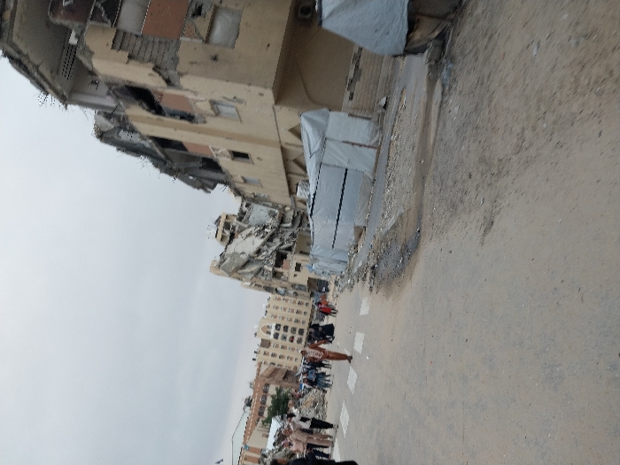 The image displays a street scene in the city of Khan Yunis, Palestinian Territory, under an overcast sky. The foreground features a dusty, uneven paved road showing scattered debris and dark stains, with a partial white pedestrian crossing visible on the left. Approximately 15-20 individuals, including men and women in civilian clothing, some wearing head coverings, walk along the left side of the street, moving away from the viewer. One person in a light brown outfit walks centrally in the mid-ground. On the right, multiple multi-story buildings are severely damaged, exhibiting partial collapse, exposed rebar, and shattered concrete facades. Adjacent to these structures, several makeshift shelters constructed from white or light-colored tarpaulins line the roadside. Extensive rubble and construction debris are present throughout the scene, particularly near the damaged buildings and shelters.