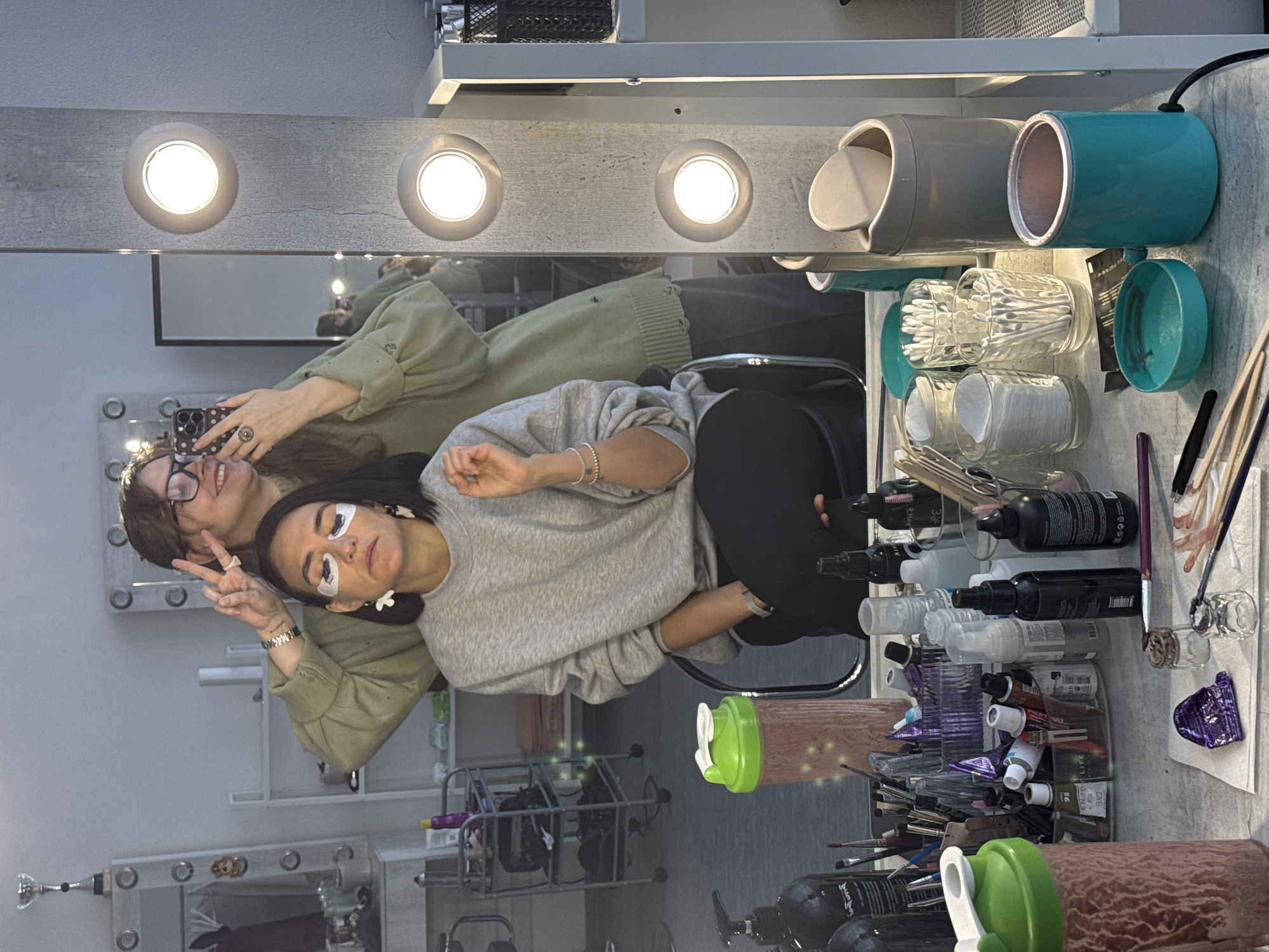 Two women are posing for a picture in front of a makeup mirror with lights, which is reflecting them. The woman in the foreground has eye patches on and is wearing a grey sweater, while the other woman is wearing glasses and a green sweater and making a peace sign. The makeup table is cluttered with various makeup products, including bottles, brushes, and containers. The reflection shows shelving units and salon equipment in the background. The location is implied to be in Izhevsk, Russia, based on external information not present in the image.
