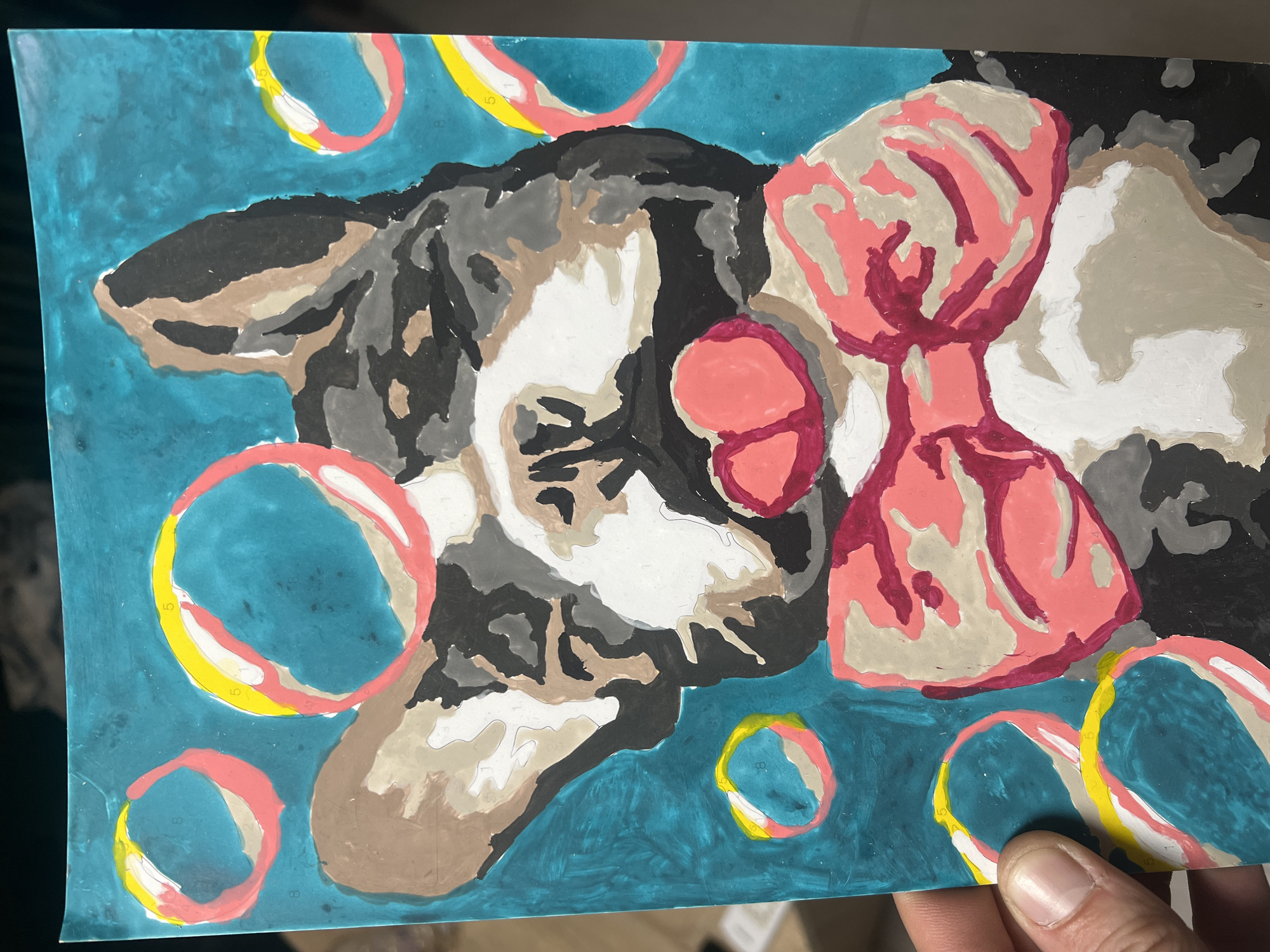 The image shows a partially completed paint-by-numbers artwork. The subject appears to be a dog wearing a bow tie, surrounded by circular bubble-like shapes. The background is a teal or turquoise color. A hand is holding the artwork in the lower right corner. There is no indication of the location as Sofia, Bulgaria.
