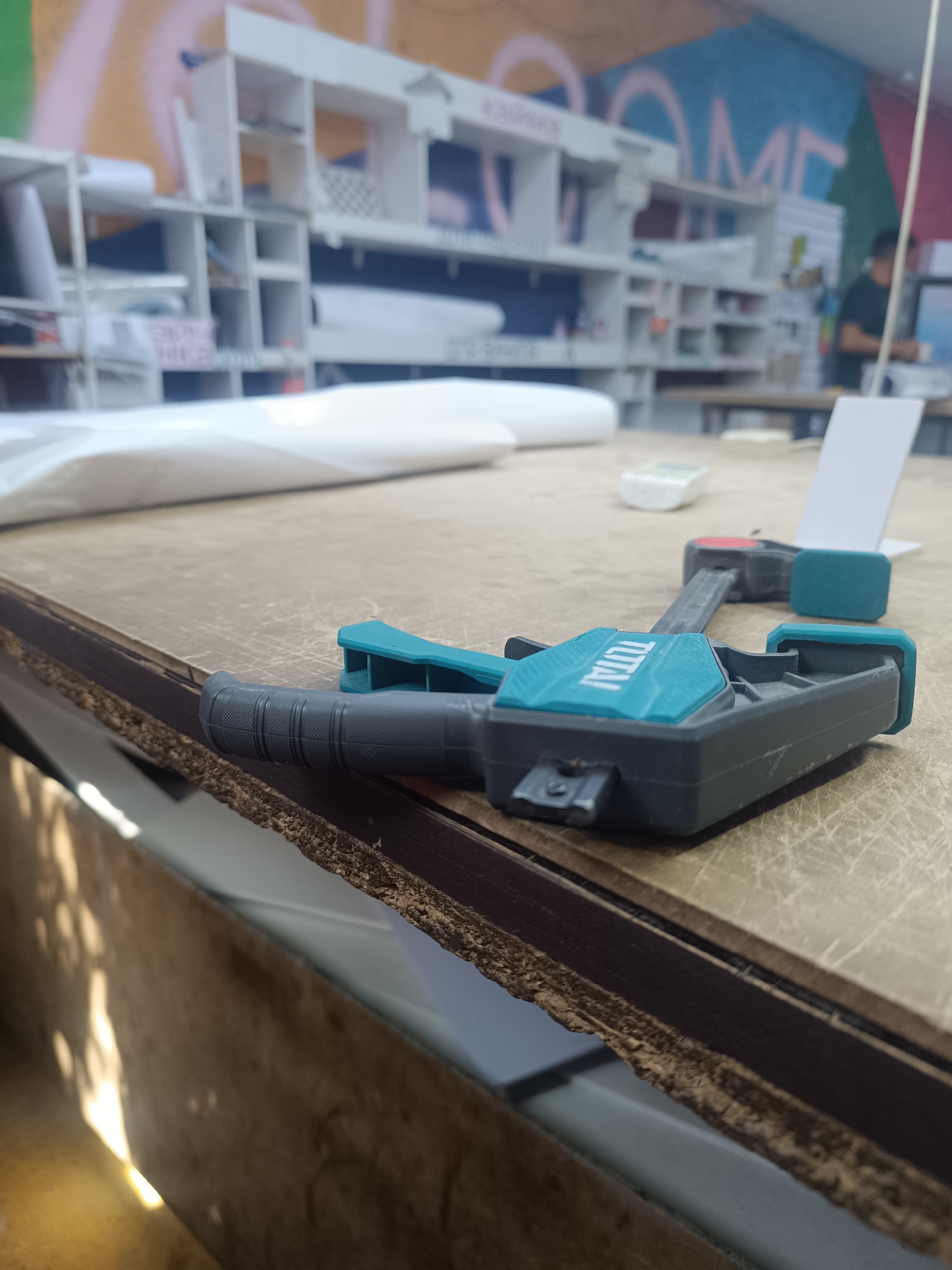 A close-up, low-angle shot focuses on a clamp resting on a wooden surface. The clamp is teal and black, with the word "TOTAL" embossed in white on its teal section. In the background, a blurry shelving unit is visible, stocked with various items. The overall impression is of a workshop or storage area. The lighting suggests it might be daytime, with shadows cast on the lower left side of the frame. The environment appears to be indoors.