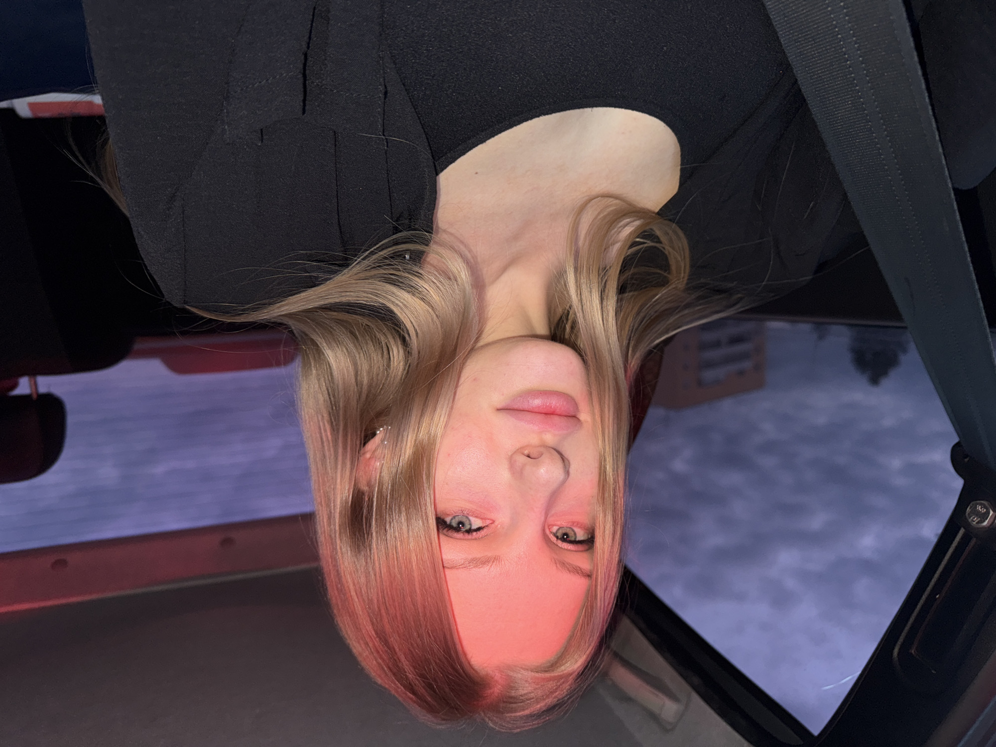 A young, fair-skinned female with blonde hair and light-colored eyes is positioned upside down in the frame. She is wearing a black top and a seatbelt is visible across her upper body. The image appears to be taken from inside a car, with the car's interior and a view of the sky through the sunroof visible. The cityscape of Perm, Russia is not discernible from the provided image.