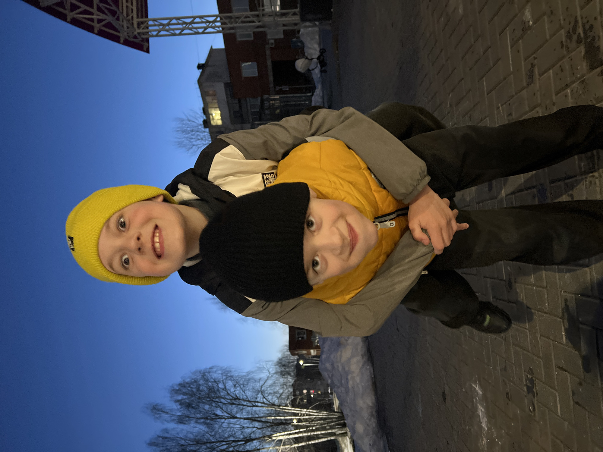 Two young boys are lying on their sides on a brick surface, possibly a sidewalk. One boy wears a yellow beanie, a black and white jacket, and dark pants. The second boy wears a black beanie and a yellow jacket. The boy in the yellow beanie is positioned on top, his arms wrapped around the other boy. They both appear to be smiling. The background shows a cityscape with buildings, trees, and patches of snow, suggesting an urban environment in Noviy, Russia. The sky is a gradient of blue, indicating twilight.