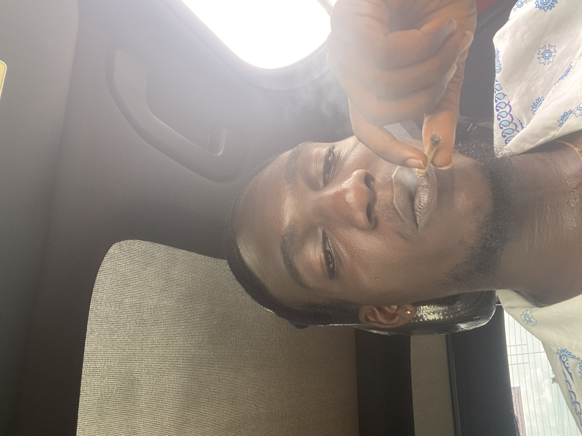 A dark-skinned male is depicted in a selfie taken from inside a vehicle, with the sun shining brightly through the car's sunroof, causing a lens flare. The man is holding a lit cigarette or blunt between his index finger and thumb, and smoke is visible around his mouth. He is wearing a white shirt with a blue paisley pattern. The background suggests an urban setting, possibly Oniyangi, Nigeria, with a blurred view of buildings through the car's rear window. The overall lighting is harsh due to the direct sunlight.