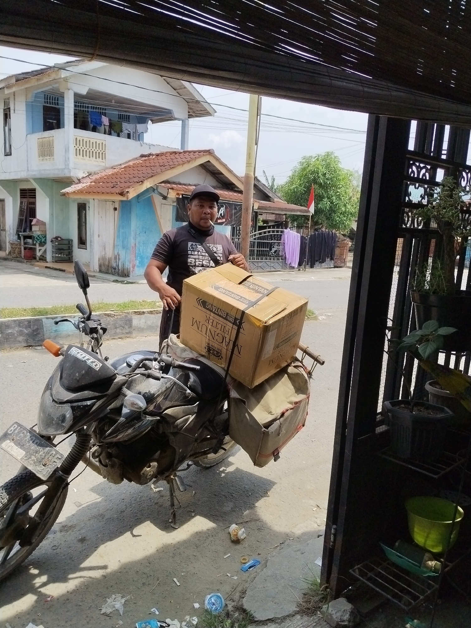 A man in a dark t-shirt and a black cap stands next to a motorcycle. He is holding a large cardboard box with text printed on it, including "MAGNUM FILTER". The motorcycle has a license plate that appears to read "BY 8269 XA". The background shows a light blue and white house with a red-tiled roof, laundry hanging on a balcony, and trees. The scene appears to be outdoors on a street with a paved road and a sidewalk. The image is taken from a slightly elevated perspective, possibly from a porch or balcony. The lighting suggests it is daytime with sunlight casting shadows. The overall impression is that of a delivery or transport scene in a residential area. The mention of Sawit Seberang, Indonesia, suggests a tropical or subtropical climate.