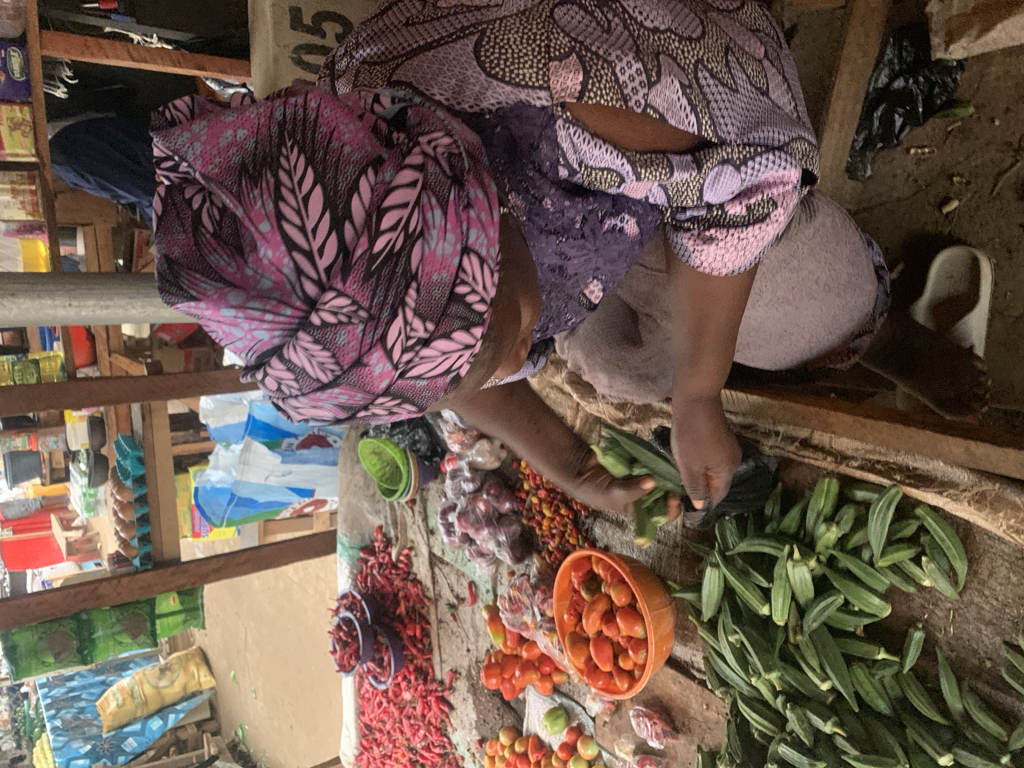 This is a vertically oriented, medium-angle, outdoor photograph of a vendor at a market in Kafanchan B, Nigeria. The vendor, a person wearing a vibrant, patterned headwrap and a purple lace top, is seated behind a display of fresh produce. In front of them are piles of red chili peppers, a bowl of tomatoes, a container of small red and yellow fruits, and a large quantity of okra. The vendor is actively selecting okra with their hands, indicating they are preparing to bag or sell it. The setting appears to be a bustling market with wooden shelves stocked with various goods in the background, suggesting a daytime scene. The ground is dirt, and there are some plastic bags and a white slipper visible. The overall impression is one of commerce and local life, with the focus on the fresh produce and the vendor's interaction with it.
