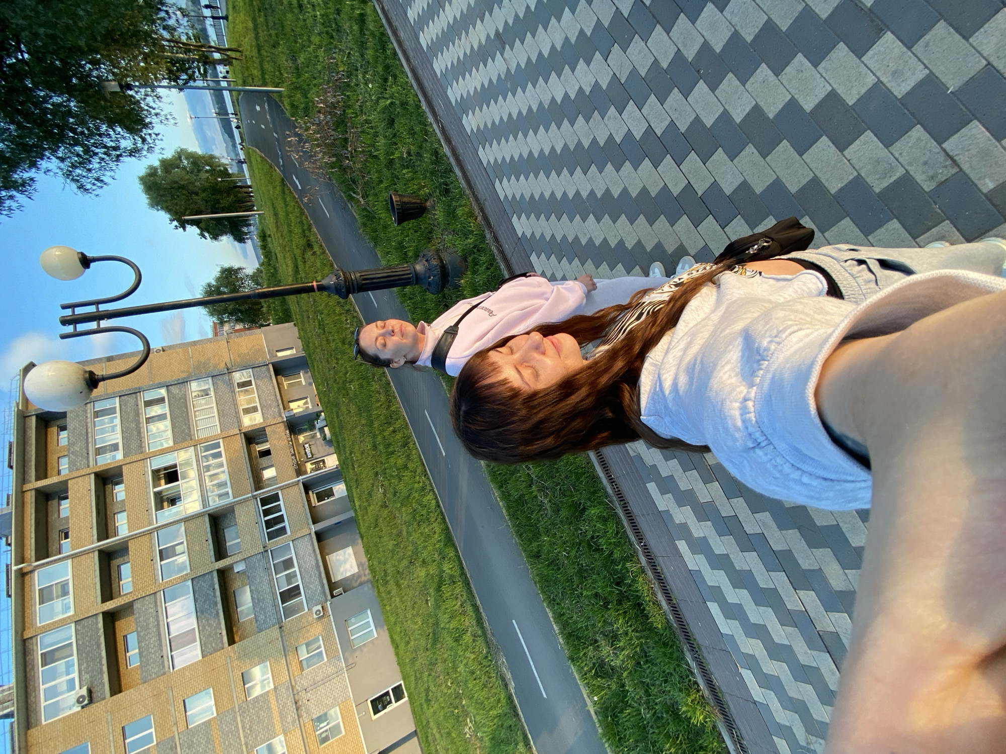The image, set in Votkinsk, Russia, captures two women outdoors during the golden hour, likely late afternoon or early evening, illuminated by warm sunlight.
In the foreground, a woman with long brown hair, wearing a white top, faces the camera. Her eyes are closed, and her face is softly lit by the sun, suggesting a moment of peace or enjoyment. Slightly behind her and to the left stands a second woman, dressed in a light pink sweatshirt and light-colored pants, with a black shoulder bag. She also has her eyes closed and a relaxed expression, seemingly basking in the sunlight.
They are positioned on a paved walkway, characterized by a distinctive diagonal pattern of alternating dark and light grey rectangular blocks. To their left, a grassy strip separates the walkway from a narrow asphalt road or bicycle path, marked with a white dashed center line. Further left stands a modern, multi-story building with a facade combining brick and concrete elements, featuring numerous windows that reflect the sky. Faint text "PHILLIPE WORLD" and "МОДА" (Russian for 'fashion') is visible on signage on the building.
Beyond the road and grassy area, a wide body of water, possibly a lake or river, stretches into the distance, with trees lining its far bank. Street lamps with double globe fixtures are positioned throughout the scene. The clear sky and low-angle sun indicate pleasant, sunny weather. The overall atmosphere is tranquil, with the women appearing to enjoy the outdoor environment.