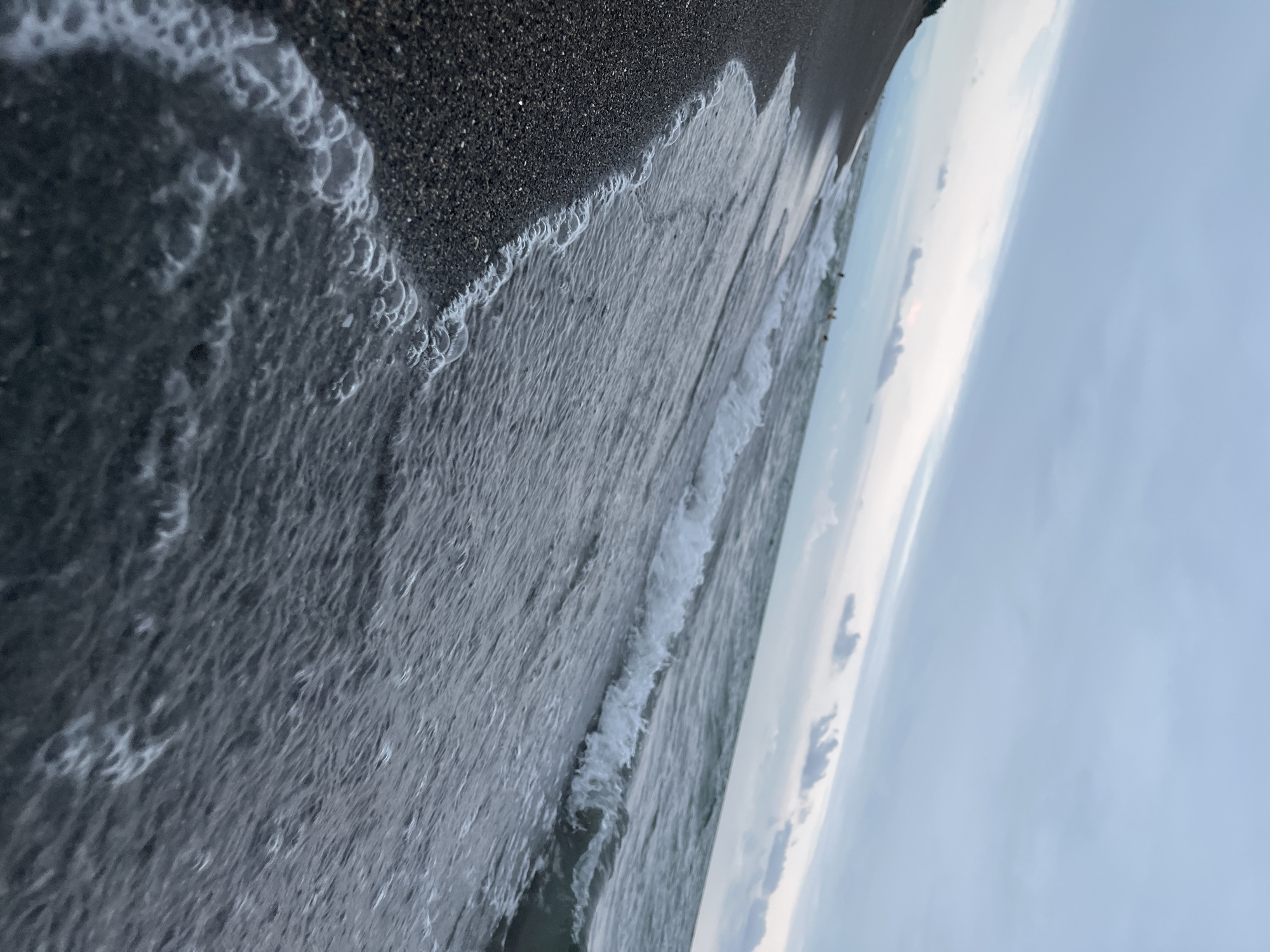 This image depicts a tranquil scene at the beach in Ureki, Georgia. The foreground is dominated by dark, grainy sand, with the edge of a gentle wave lapping at the shore. The water is a deep, muted grey, reflecting the overcast sky. Small, white bubbles and foam are visible along the waterline, testament to the waves' movement. In the distance, the sea stretches towards the horizon, where it meets a sky filled with layers of grey and pale blue clouds. A few faint, dark shapes in the water might be people swimming or wading. The overall impression is one of a calm, perhaps cool, day at the seaside.