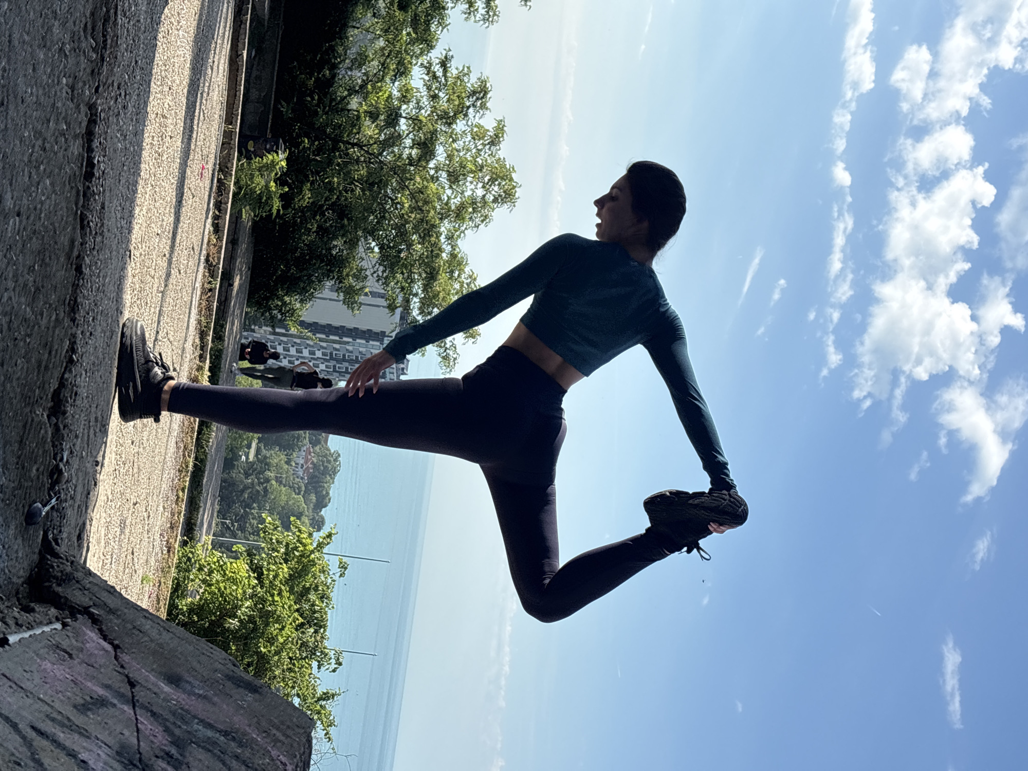 The image displays a young woman performing a stretching or yoga pose outdoors, captured in a portrait orientation that has been rotated 90 degrees clockwise. She stands on a textured concrete or stone pathway, which serves as the foreground and extends along the left side of the frame.
The woman, with dark hair tied back, is dressed in dark teal athletic crop top with long sleeves, dark leggings, and black athletic shoes. She is balancing on one leg (her right) while her left leg is bent upwards and backward, with her left hand holding her left foot, indicating a deep stretch. Her right arm is extended forward for balance, and her body is slightly twisted. Her head is turned towards the right and slightly upwards, with an open-mouthed expression that suggests concentration, exertion, or possibly a moment of breath release during the pose.
The setting is bright and sunny, indicative of daytime, likely morning or afternoon, with clear shadows cast by the sun. The weather appears pleasant, featuring a clear blue sky adorned with scattered white cumulus clouds. In the mid-ground, a wide expanse of calm blue water, identifiable as the Black Sea given the Varna, Bulgaria location, stretches towards the horizon. Lush green trees and foliage are visible both near the woman and in the distance, interspersed with distant buildings or structures that imply an urban or semi-urban coastal environment. The overall impression is one of fitness and well-being amidst a scenic natural landscape. There is no visible text in the image.