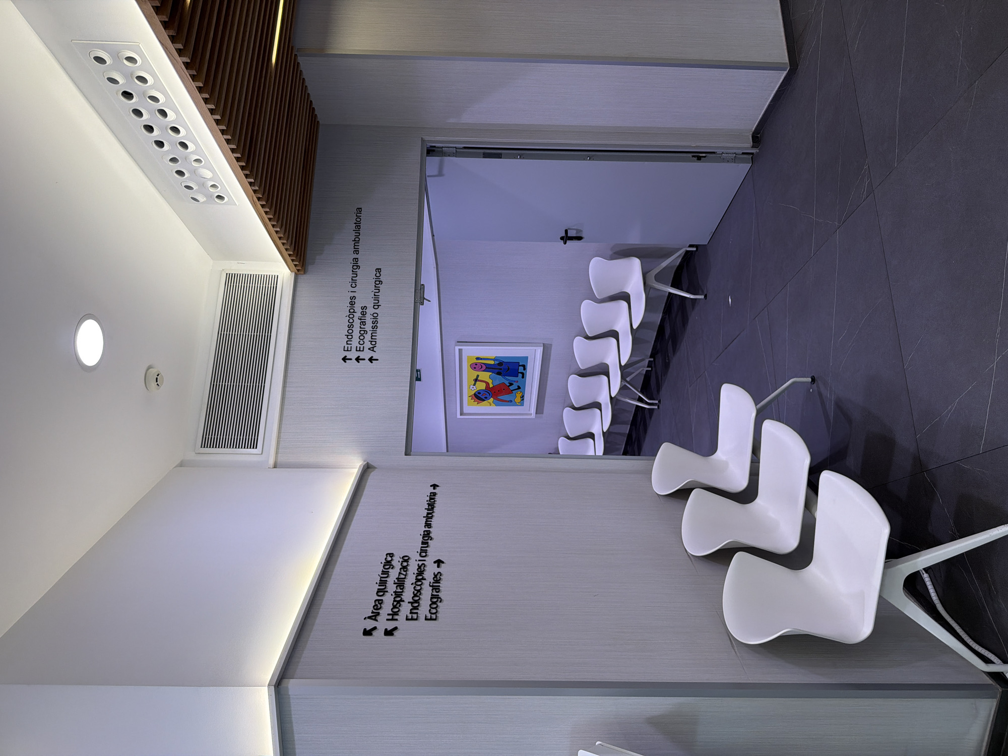 This is a wide-angle, eye-level, indoor photograph of a modern clinic waiting area in Palma de Mallorca, Spain. The image captures a sterile, minimalist environment with a focus on white furniture and muted grey walls.
In the foreground, three white, molded plastic chairs with thin metal legs are arranged against a grey textured wall. The chairs have a distinctive curved back and seat. The floor is covered with large, dark grey tiles that have a subtle white veining pattern.
On the left wall, there are two sets of directional signs with black lettering and symbols. The upper sign, pointing left, reads "Àrea quirúrgica" (Surgical Area). Below it, also pointing left, is "Hospitalització" (Hospitalization). To the right of these, a sign points forward and reads "Endoscòpies i cirurgia ambulatòria" (Endoscopies and ambulatory surgery). Below this, another sign points forward and reads "Ecografies" (Ultrasounds).
On the right side of the image, an open doorway leads into another room. Above the doorway, a similar set of directional signs are mounted on the wall, pointing upwards: "Endoscòpies i cirurgia ambulatoria", "Ecografies", and "Admissió quirúrgica" (Surgical Admission). Inside the room visible through the doorway, there are more white chairs and a framed piece of colorful abstract art on the wall.
The ceiling features recessed lighting and ventilation grilles. A distinct wooden slatted panel runs along the upper right portion of the ceiling. The overall impression is one of cleanliness, efficiency, and a modern healthcare facility. There are no people visible in the photograph.