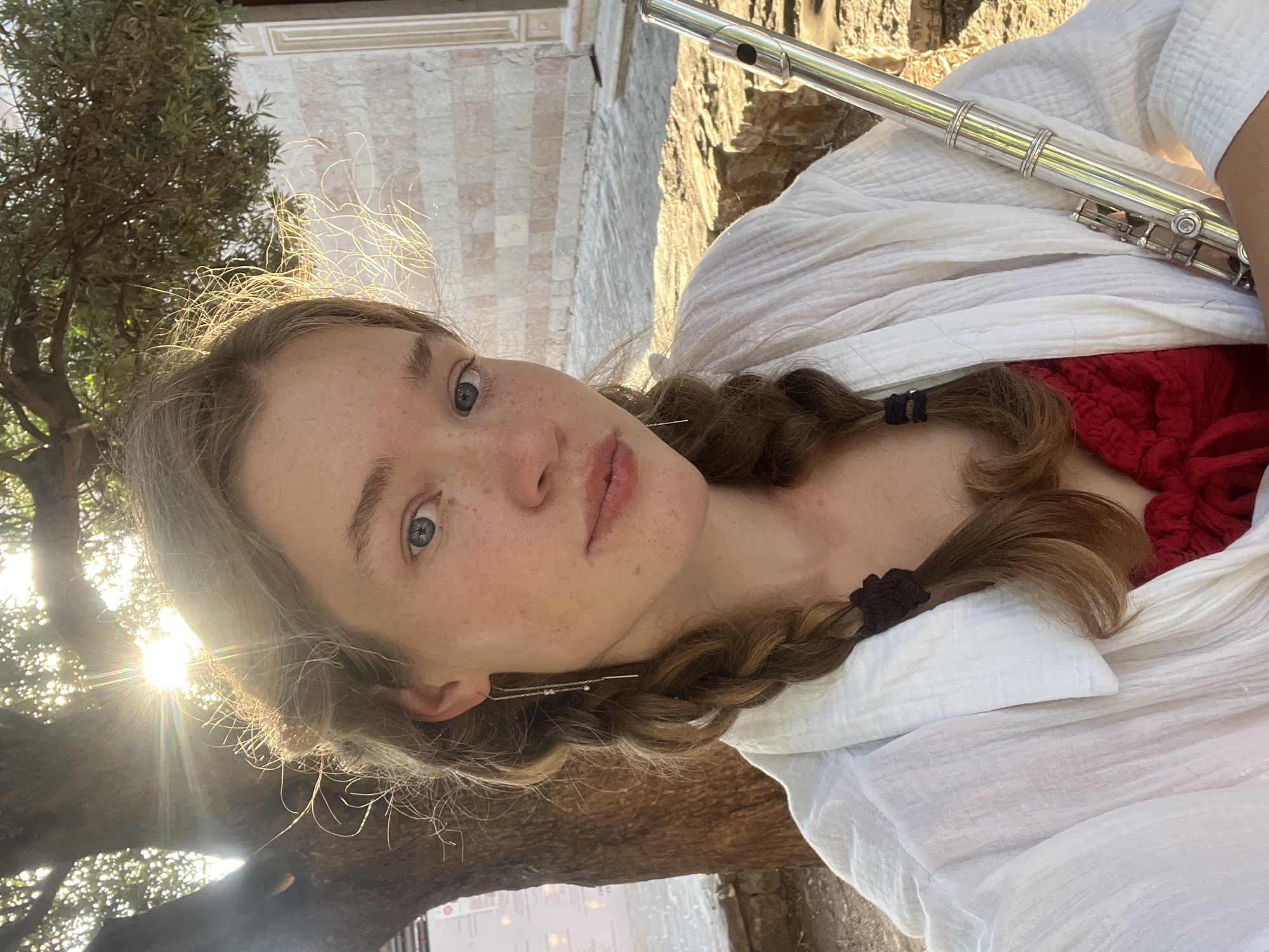 A fair-skinned woman with light brown hair styled in two braids, secured with dark-colored hair ties, is gazing towards the camera. She has freckles and light-colored eyes. She's dressed in a red top covered by a white, textured shawl or wrap. Part of a silver flute is visible, held by the woman but partially out of frame. The photo appears to be taken outdoors in a sunlit environment. To the left, a tree with green foliage is partly visible, with the sun shining through its branches. In the background, there's a structure with light-colored stonework. Based on the style of the stonework, the tree, and the overall ambience, it is plausible that the location is Budva, Montenegro.