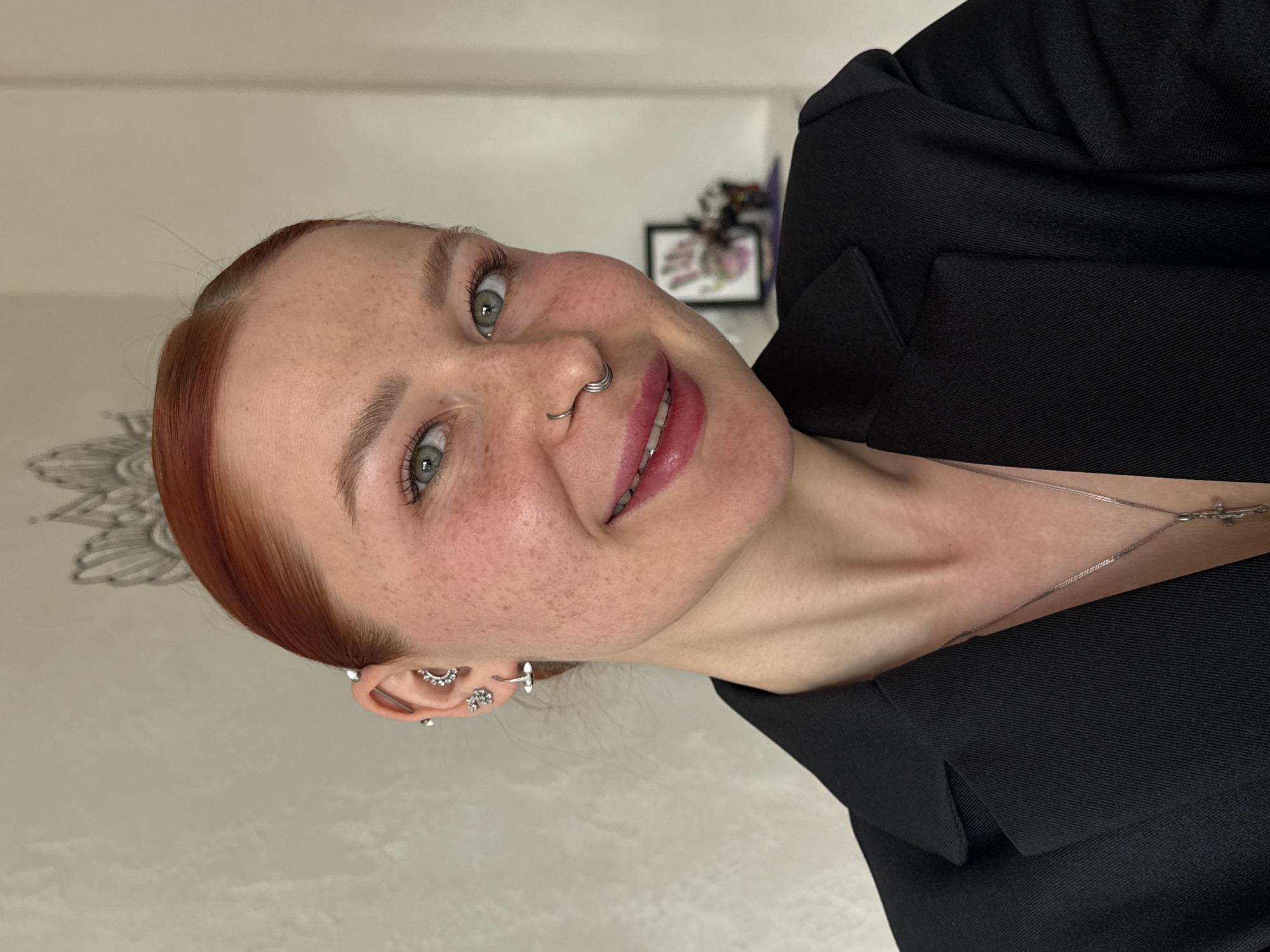 A close-up photograph shows a fair-skinned woman with red hair, freckles, green eyes, and multiple piercings in her left ear and nose. She is smiling slightly. She wears a black blazer and a silver necklace with a cross pendant. The background is a blurred white wall with a faint decorative pattern. The location is not identifiable as Rostov-on-Don, Russia, from the image.