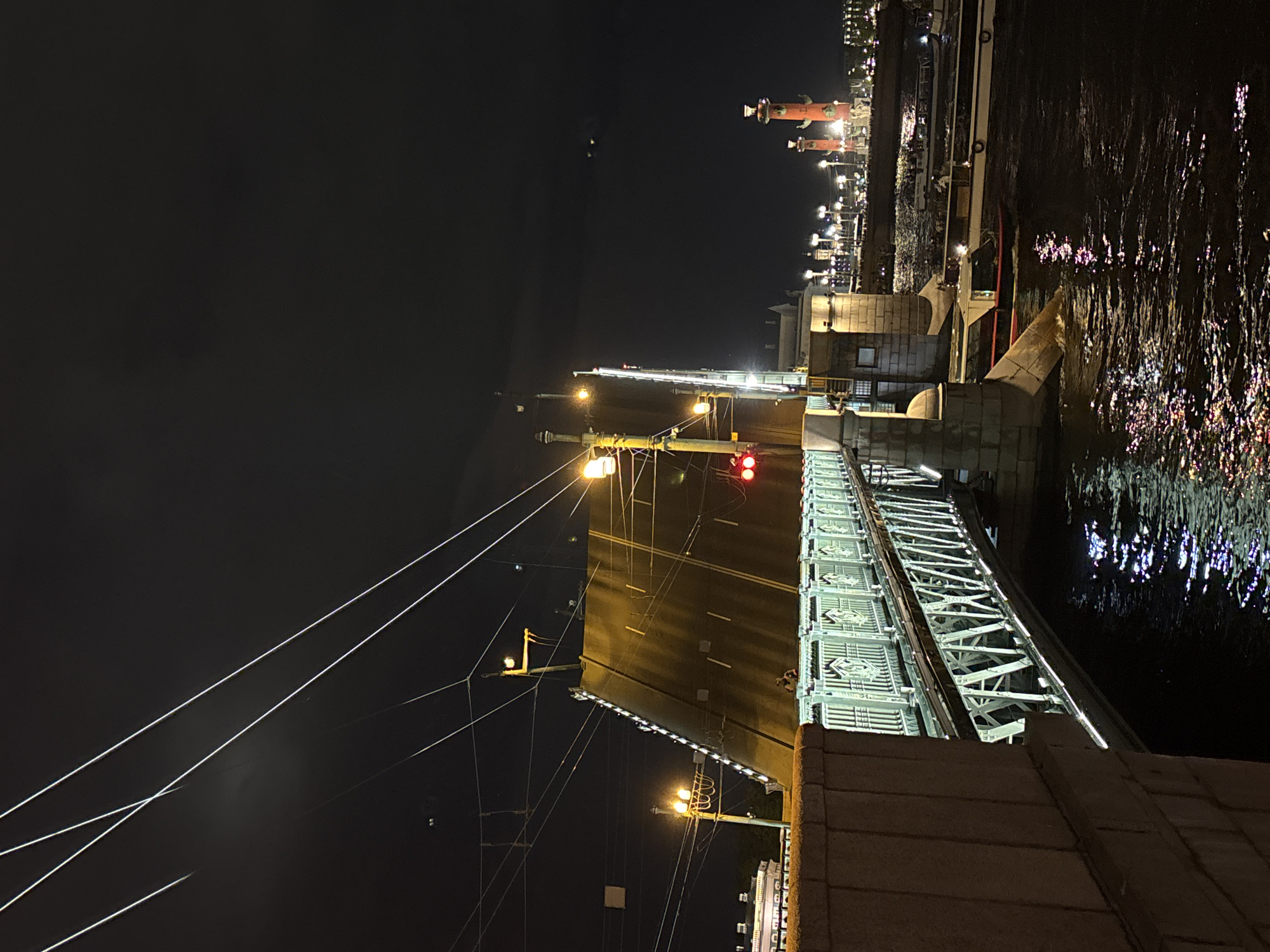 At nighttime, the image depicts the Palace Bridge in Saint Petersburg, Russia. The bridge, a key landmark, spans the Neva River, illuminated by street lights. The Rostral Columns can be seen in the background along the river. The reflective surface of the river shows light patterns. The bridge structure is visible, extending over the water and connecting different parts of the city. The overall atmosphere is that of a city at night, with architectural and urban features dominating the scene.