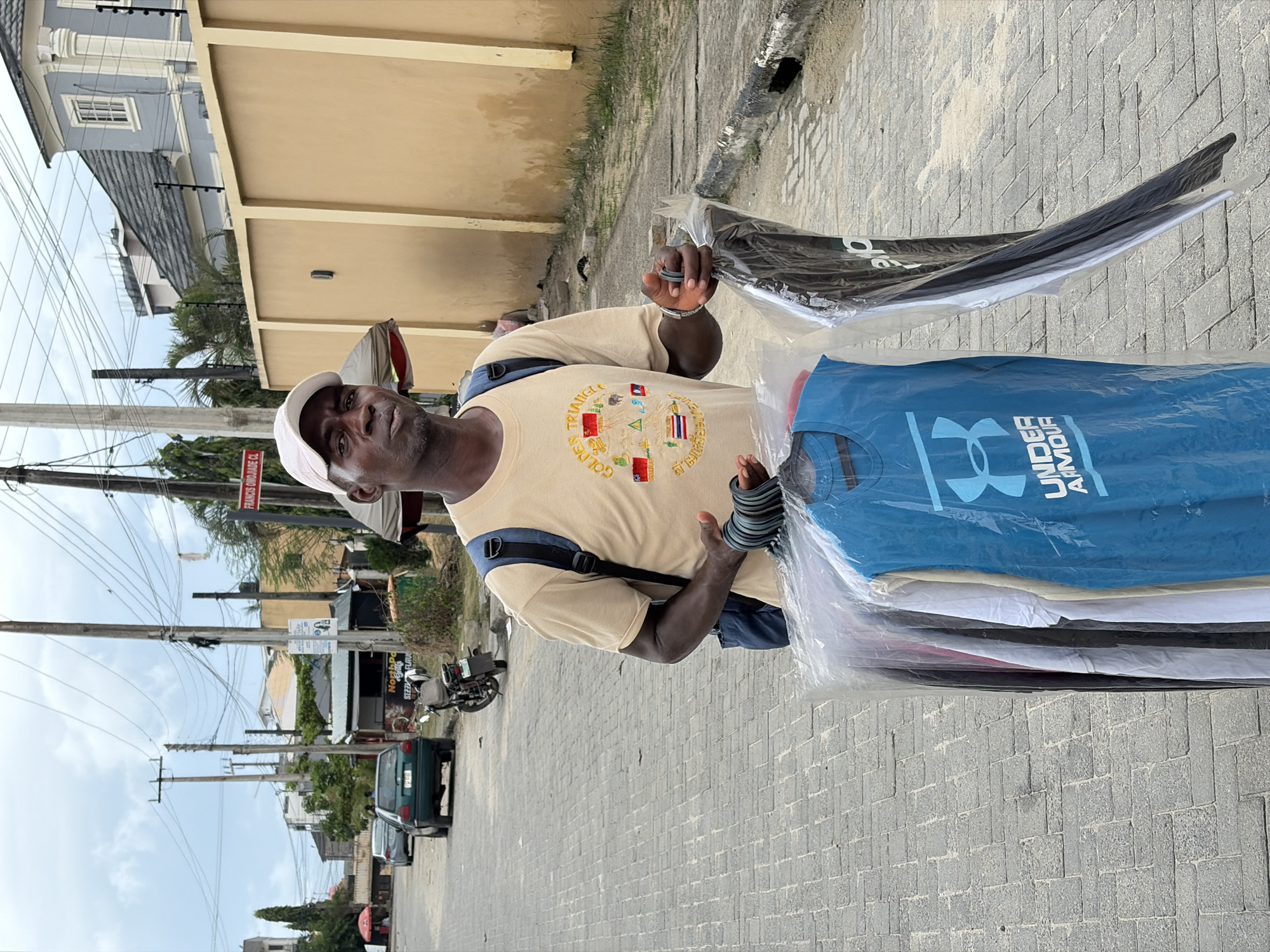 A man, appearing to be in his late 40s or 50s, stands outdoors holding a stack of folded t-shirts wrapped in clear plastic. He wears a light tan t-shirt with a circular emblem on the chest featuring flags and geometric shapes, and a white baseball cap. His left arm is in a blue sling. The t-shirts in his hand are predominantly black, white, and blue, with the blue one prominently displaying the "Under Armour" logo.
The man is on a paved street made of grey bricks. Behind him, a beige wall runs along the side of a building. Further in the background, a modern, multi-story building with grey siding and white trim is visible, along with power lines and utility poles. The scene suggests an urban or suburban environment, likely in Igboefon, Nigeria, given the context.
The lighting suggests it is daytime, with a bright sky, although the orientation of the image makes it difficult to ascertain the exact time or specific weather conditions. The man's expression is neutral. The presence of a parked car and a motorcycle in the background further indicates a street scene. Visible text includes "UNDER ARMOUR" on the blue t-shirt and a partial sign on a pole in the background that reads "FRANKS FOOD AND".