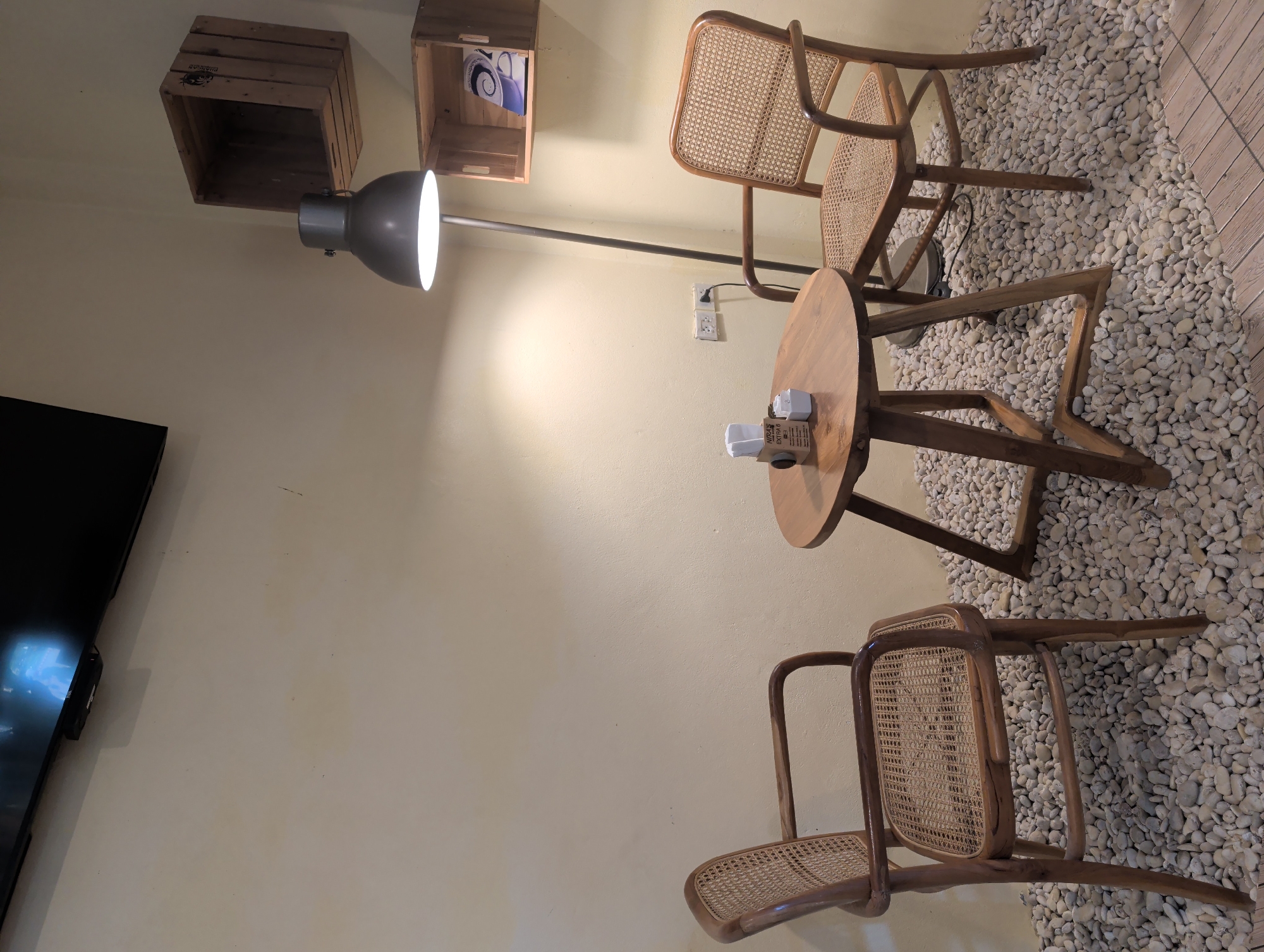 This interior scene features a small seating arrangement positioned on a floor covered in small, white pebbles. The arrangement consists of two wooden chairs with woven backs and seats, flanking a small, round wooden table. On the table, there are items that appear to be disposable coffee cups or containers.
A floor lamp with a gray, dome-shaped head is positioned behind the table and chairs, providing illumination. The scene is set against a light beige wall. Wall-mounted wooden crates are positioned higher on the wall. The environment appears to be an indoor space, and the style suggests a cafe or lounge. Based on the prompt, the location is identified as Thong Sala, Thailand, but there is no direct evidence of this in the image. A television screen is visible on the left.