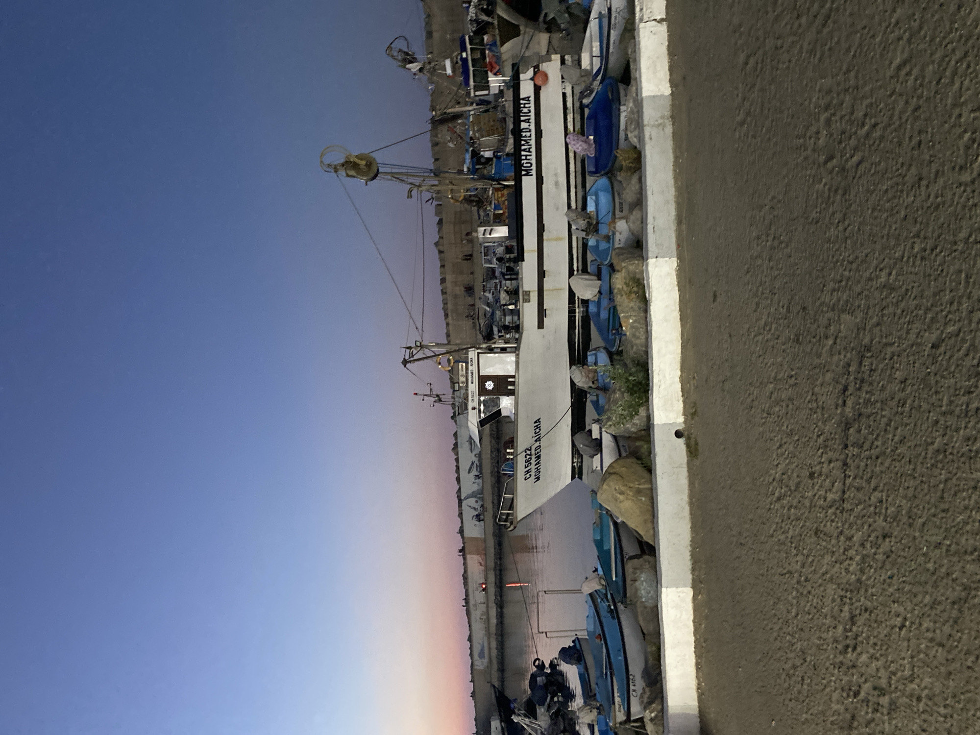 A fishing boat named "MOHAMED AICHA" with the registration "CH 5622" is docked in a harbor at dusk, likely in Tipaza, Algeria. Several smaller blue and white boats are moored alongside it. In the background, a concrete wall with scattered figures can be seen, suggesting a populated area. The sky is a gradient of blue and pink, indicating the transition from day to night. The foreground is a sandy embankment.