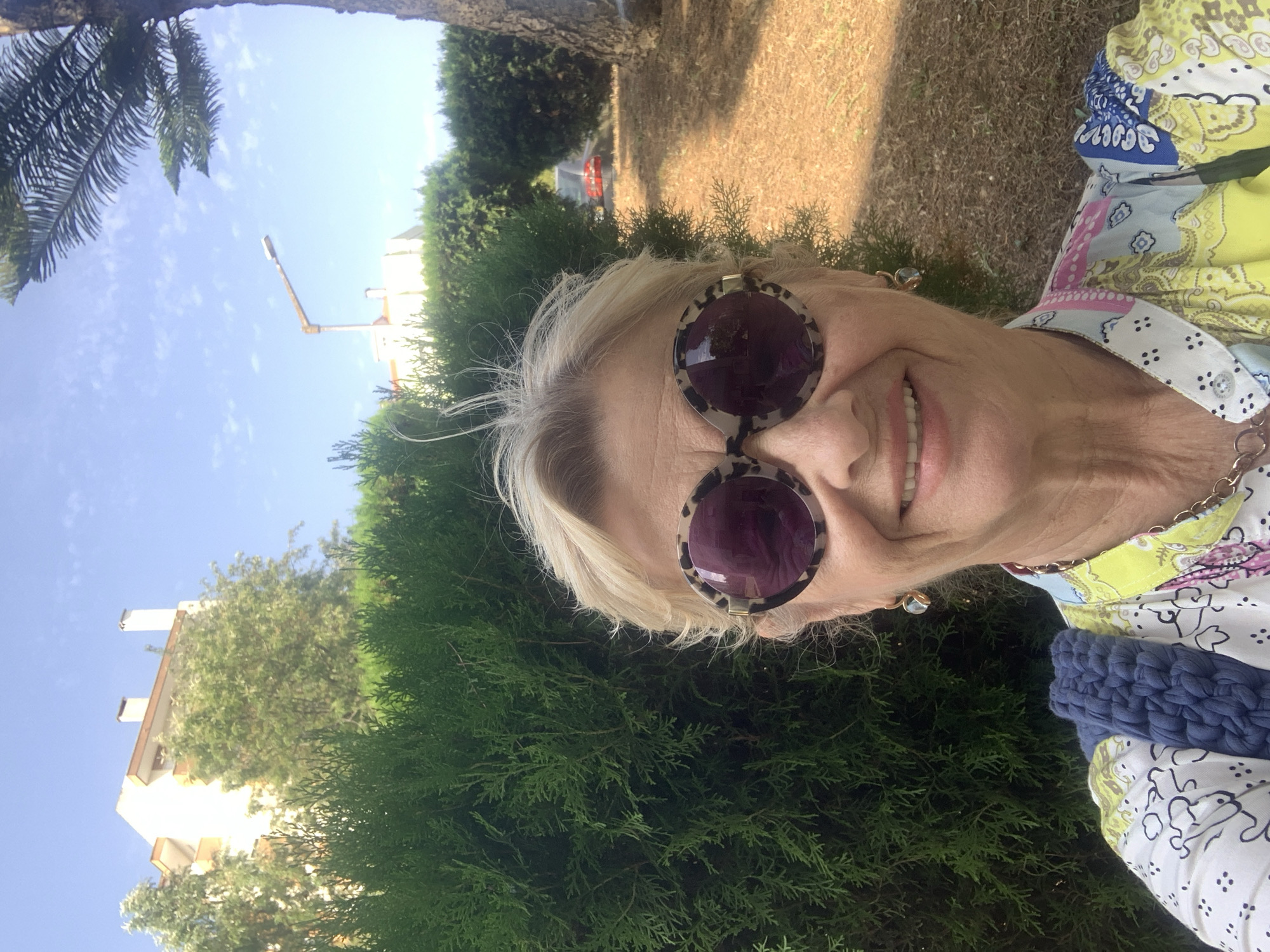 This is a selfie taken outdoors in Odivelas, Portugal, likely during the daytime given the bright sunlight and clear blue sky. The subject is a woman with blonde hair, wearing large, round sunglasses with a leopard-print frame. She is smiling, and her face is turned towards the camera. She is wearing a colorful, patterned shirt with blue, yellow, and pink elements. A navy blue, textured accessory, possibly a bag or scarf, is visible on her right side.
The background consists of lush green foliage, including dense bushes and trees, and a patch of dry grass and dirt. In the distance, a building with a cream-colored facade and chimneys can be seen, along with a street light and a hint of a palm tree on the left side of the frame. The overall impression is of a sunny day in a suburban or park-like setting. The image is oriented vertically, as if the camera was held in portrait mode.