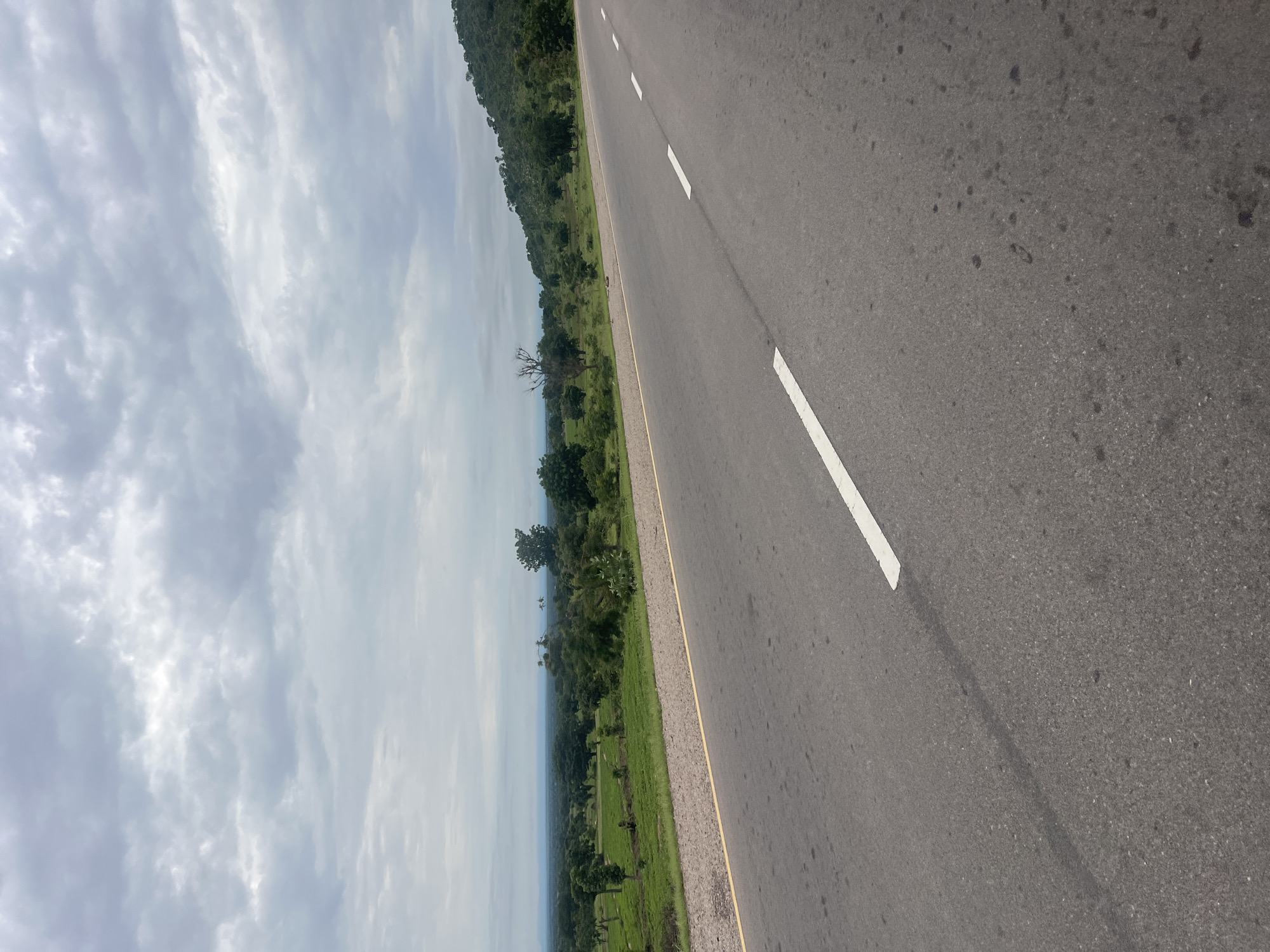 The image shows a paved road with white dashed lines, stretching towards a landscape of green fields, scattered trees, and a distant hazy horizon. A gentle slope rises on the right side of the road, covered with trees. The sky above is overcast with dense, gray clouds. The road surface appears to be asphalt, with some darker patches and a yellow line marking the shoulder on the left. The overall impression is one of a rural or semi-rural environment under a cloudy sky, possibly during the wet season. The location is stated to be in Minda, Nigeria.