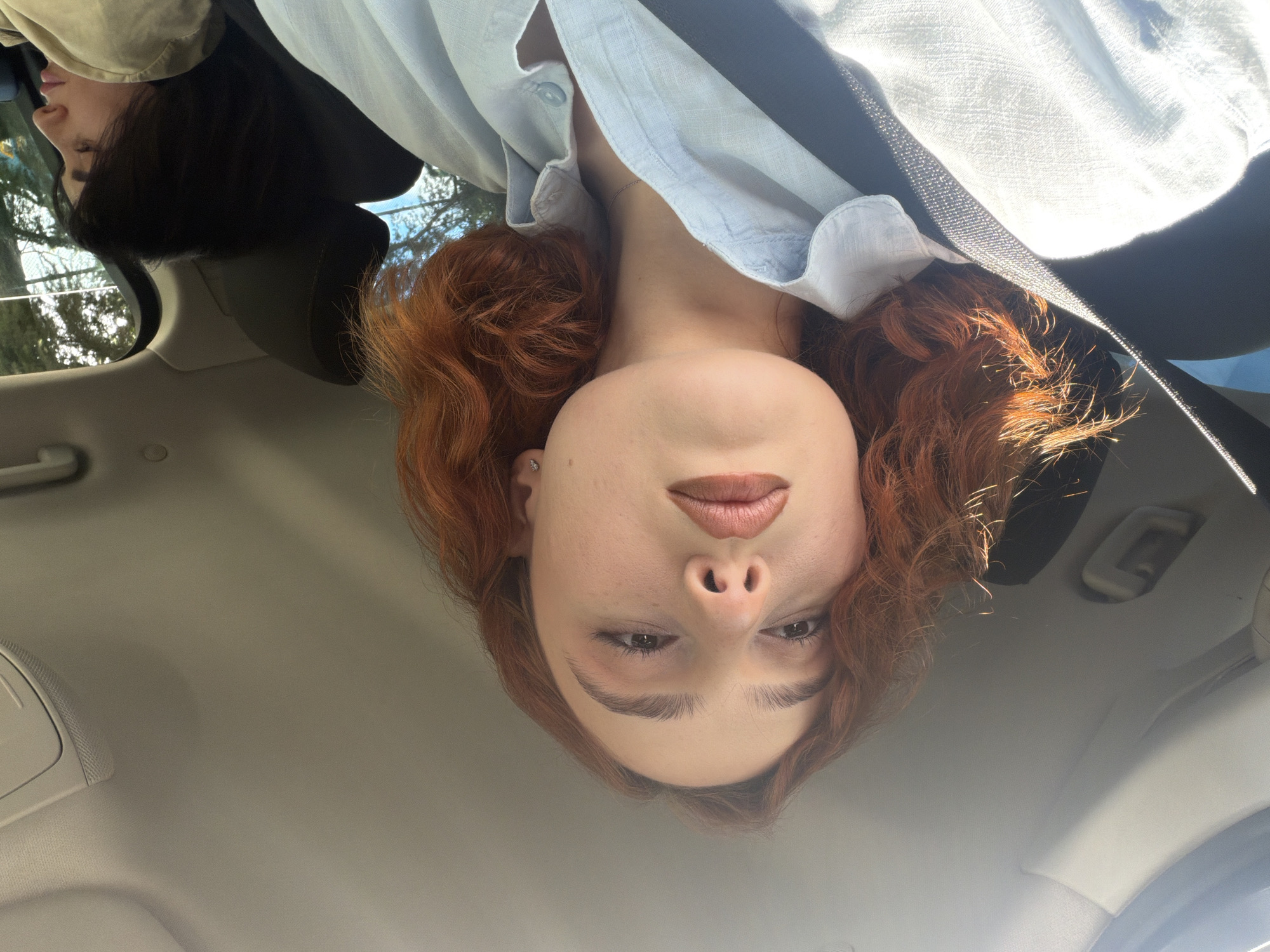 A close-up, inverted indoor shot taken in a vehicle shows two individuals. One with dark hair is partially visible in the upper left corner, while the other is facing the camera directly. This person has fair skin, red curly hair, brown eyes, and full lips. They are wearing a light blue button-up shirt and a seatbelt. The interior of the car includes a light gray headliner and various car fixtures. There is no verifiable evidence in the image to identify the location as Tbilisi, Georgia.