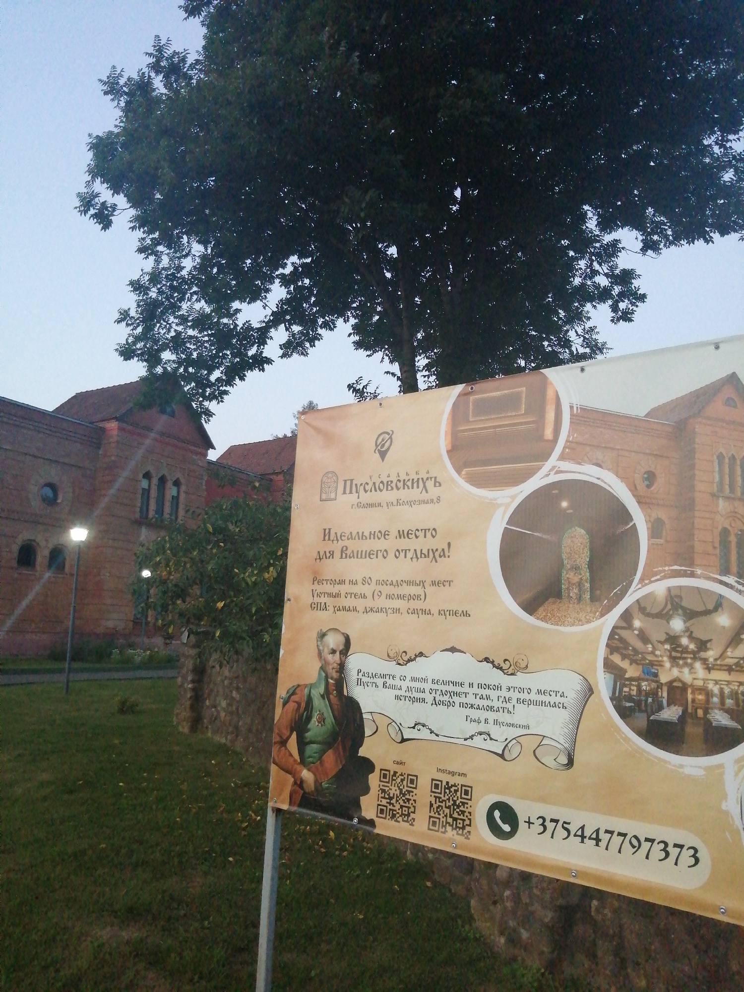 The image shows a banner with information about "Usad'ba Puslovskikh" (Puslovskikh Estate) located in Slonim, Belarus. The banner features images of the estate, an interior view of a restaurant, and a portrait of a man. Text on the banner advertises it as an "Ideal place for your vacation," and provides details about the restaurant's capacity, accommodation options, and amenities such as a sauna and a pool. Contact information, including a phone number and QR codes for a website and Instagram, is also displayed. In the background, a brick building with a red roof, likely part of the estate, is visible, along with trees and a grassy area. The overall scene suggests an advertisement for a historical and recreational complex.