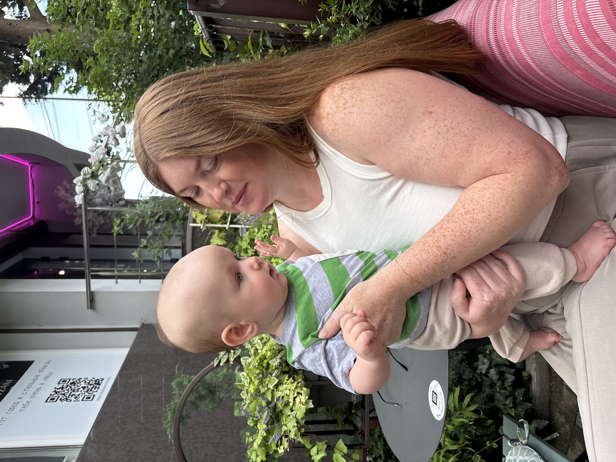 The image shows a woman holding a baby outdoors. The woman has long, reddish-brown hair and fair skin with freckles. She is wearing a white tank top. The baby is wearing a striped green and gray shirt and beige pants. The baby is looking up at the woman. They are sitting in a setting that appears to be a cafe or outdoor seating area, with lush greenery and trees in the background. There are also some architectural elements, including a building with a pink neon sign.
The woman appears to be smiling gently at the baby, and the baby has an engaged expression. The lighting suggests it is daytime, possibly late afternoon, with soft natural light. The presence of greenery and the overall atmosphere indicate a pleasant outdoor environment.
On a table in the foreground, there is a circular sticker with a QR code and some text. Although the text is mostly illegible in the provided image, some Cyrillic characters can be seen, suggesting a Russian context. Given the location context of Sochi, Russia, the scene likely depicts a relaxed moment in a public outdoor space in that city.