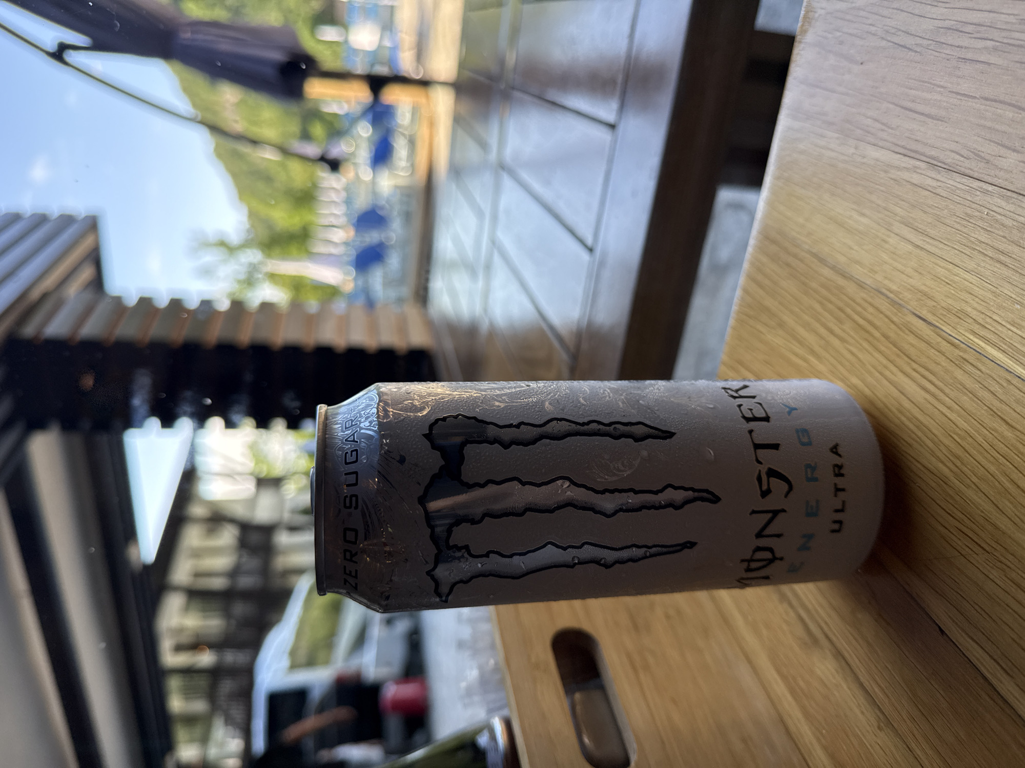 A close-up shot depicts a can of "Monster Energy Ultra" drink lying on a wooden surface. The can, adorned with the Monster claw logo, is partially covered in condensation. In the blurred background, there's a building with a partially visible awning. Through the structure, a glimpse of what appears to be a mountain or a park setting can be discerned, suggesting an outdoor location. Given the prompt mentions Veliko Tarnovo, Bulgaria, the scene could be a restaurant or café with outdoor seating in this city.