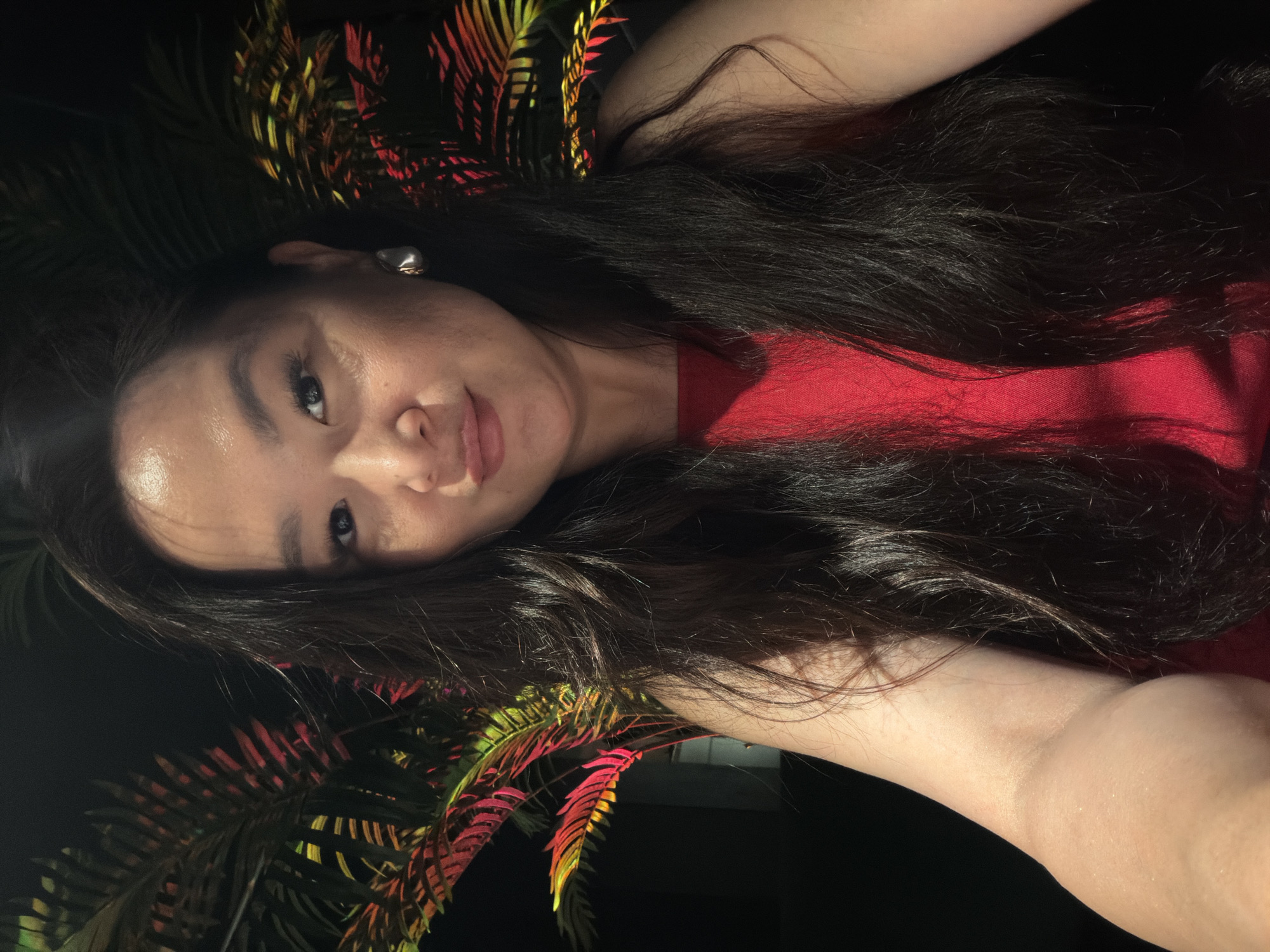 A woman with long, dark hair, wearing a red top and a silver earring, is posing for a selfie with tropical foliage in the background. The location is difficult to determine, but based on the prompt, it is somewhere in the city of Chaikovsky, Russia. The lighting is dim, suggesting nighttime or an indoor setting with limited light.