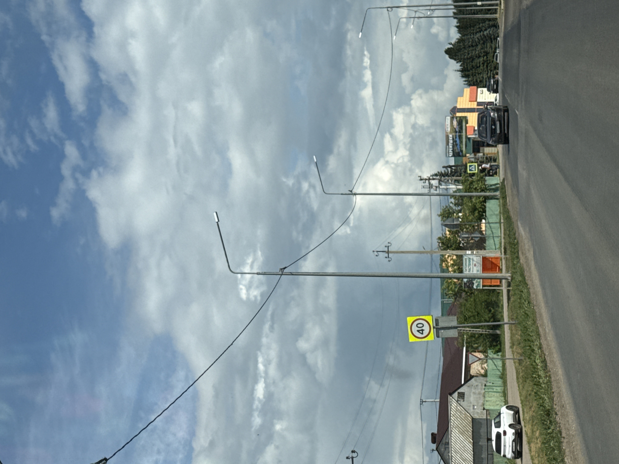 This image depicts a street scene in Nurykeevo, Russia. A paved road runs from the foreground towards the background, bordered by grassy verges and a green wooden fence on the left. Several streetlights are visible along the road, with overhead wires connecting them. A white SUV is parked on the left side of the road. Further down the road on the left, there are some residential buildings with pitched roofs. A yellow speed limit sign indicating "40" is mounted on a pole. On the right side of the road, a colorful building with signs is visible, and a line of coniferous trees stands behind it. Two cars are driving on the road in the distance. The sky is partially cloudy.