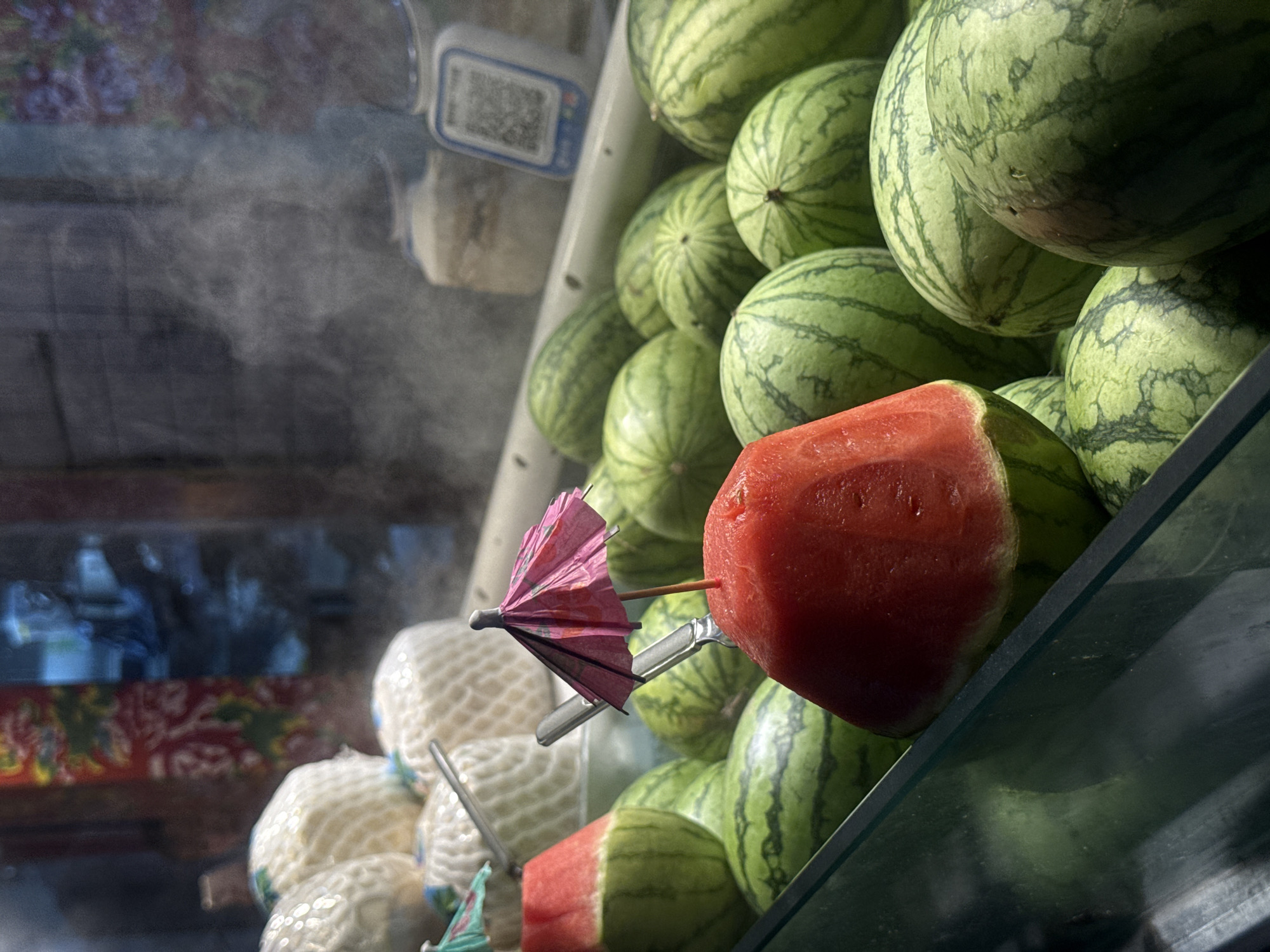 This image depicts a close-up view of a display of fresh watermelons, some of which are cut to reveal their red flesh. Several of the whole watermelons have dark green stripes on their rinds. A small, pink paper umbrella is placed on top of a cut piece of watermelon. In the background, there are stacked white netted items, possibly coconuts, and a QR code scanner is visible. A faint mist or steam rises from the display. The overall scene suggests a market stall or fruit vendor, possibly in Qinhuangdao, China, where such vibrant fruit displays are common. The lighting is somewhat dim, with the focus primarily on the fruit.