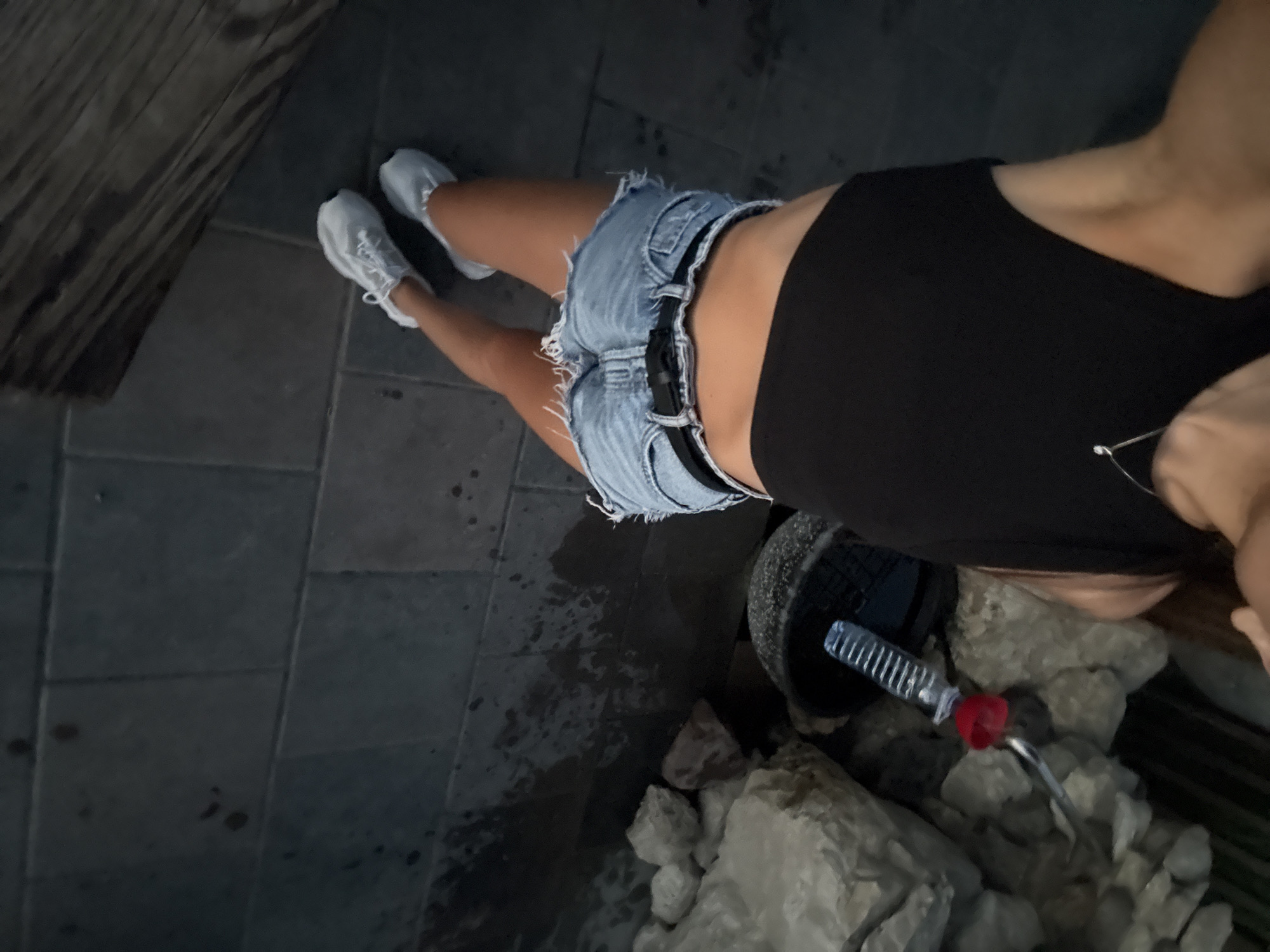 The image shows a person wearing a black crop top and denim shorts, with white sneakers. The person is positioned on a paved surface, possibly a sidewalk or patio, with a wooden structure to the right. In the background, there are large rocks and a black bucket containing a plastic bottle with a red lid and a metal straw. The scene appears to be outdoors, and the lighting suggests it may be evening or a dimly lit area. No specific details indicate the location in Cimișlia, Moldova.