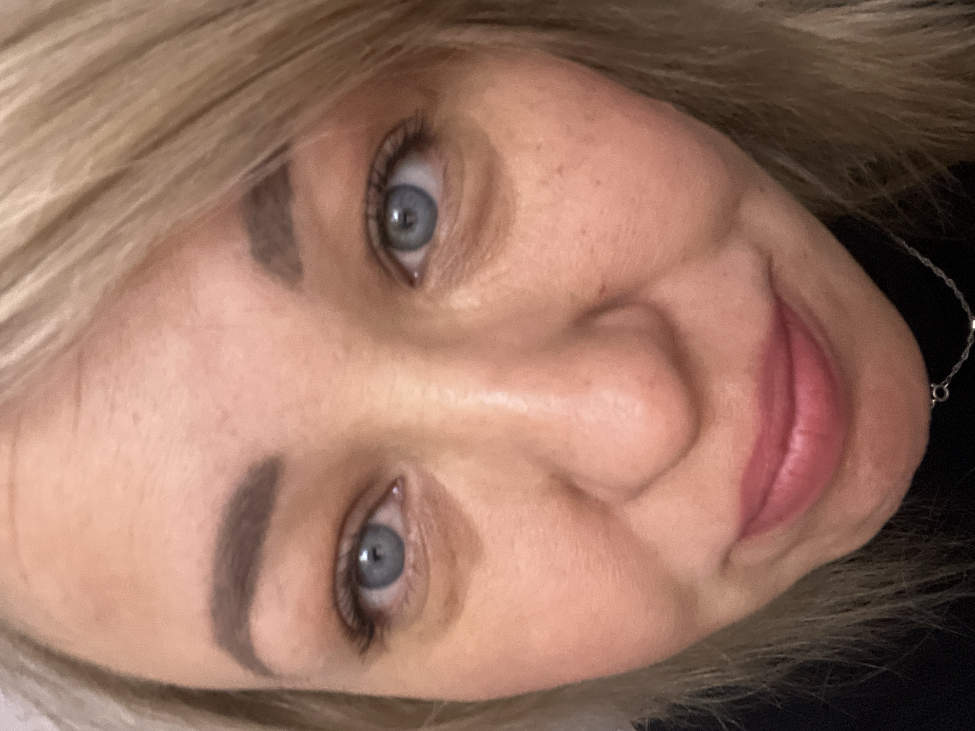 A close-up, slightly tilted portrait of a fair-skinned woman with blonde hair and blue eyes. Her face is visible from the forehead to the chin, and includes eyebrows, nose, and lips. She has fair skin with some freckles visible on her nose and cheek. The background is dark, but a hint of a necklace can be seen. There is no information provided in the image to suggest the location is Chaikovsky, Russia.