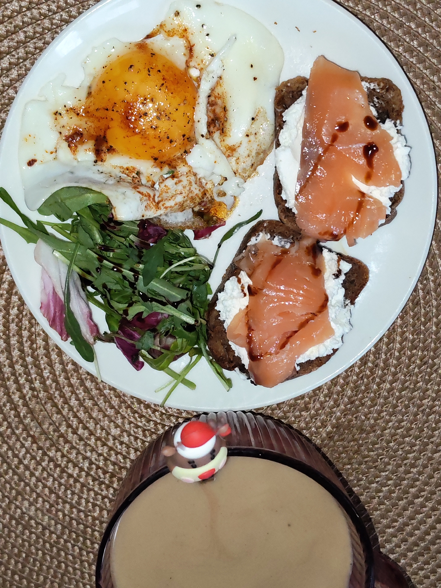 This overhead shot captures a visually appealing meal arranged on a white plate, accompanied by a beverage in a glass. The scene is set indoors, likely at a home or restaurant in Izhevsk, Russia, with the dishes resting on a light brown, woven placemat.
The main plate features a breakfast or brunch spread. On the left are two fried eggs, one with a distinct, golden-orange yolk that appears slightly runny, both seasoned with black pepper and a reddish spice. Adjacent to the eggs is a small serving of fresh mixed greens, including arugula and radicchio, dressed with a dark, glossy balsamic glaze or similar dressing. To the right, there are two open-faced bites or small toasts, made from dark, possibly whole-grain or rye bread. Each is topped with a generous dollop of white, creamy cheese (such as ricotta or cream cheese) and layered with slices of delicate, light orange smoked salmon, also adorned with a drizzle of the dark glaze.
Partially visible in the lower section of the frame is a glass containing a light brown beverage, likely coffee or a coffee-based drink. A small, festive decoration, appearing to be a marshmallow or candy shaped like a reindeer wearing a red Santa hat, is perched on the rim of the glass. No people are visible, and there is no discernible text in the image. The overall presentation suggests a well-prepared and enjoyable meal.