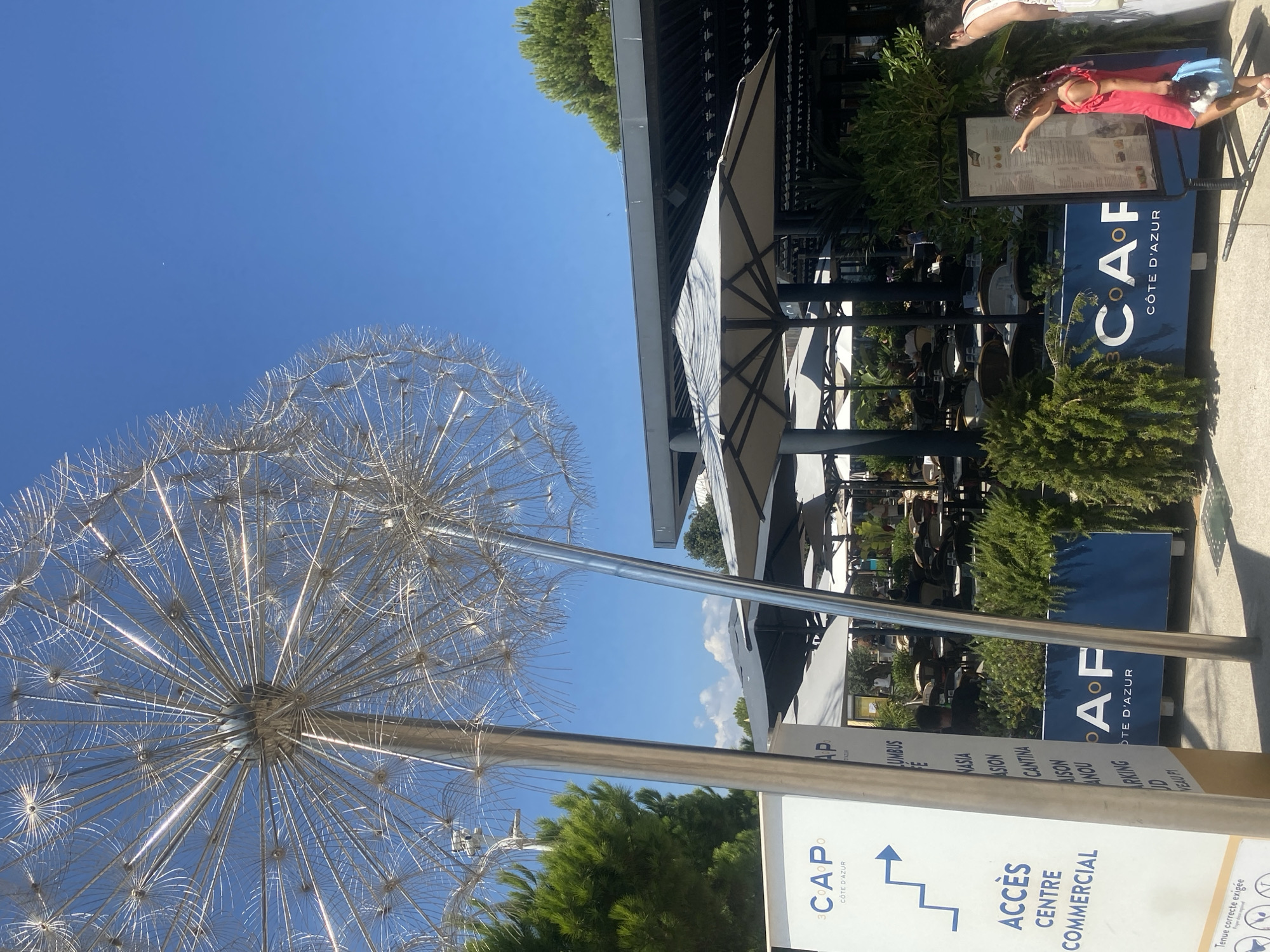 The image depicts an outdoor seating area of a restaurant or cafe, identified by signage as "CAP CÔTE D'AZUR" in Saint-Laurent-du-Var, France. Two large, metallic dandelion sculptures are prominently featured in the foreground. The seating area, partially covered by grey umbrellas, is visible in the background with tables and chairs set up. Green foliage and trees surround the area. A white sign with blue text and an arrow indicates "ACCÈS CENTRE COMMERCIAL." In the lower right, a young girl in a pink dress and a woman are looking at a menu board. The scene is set under a clear blue sky.