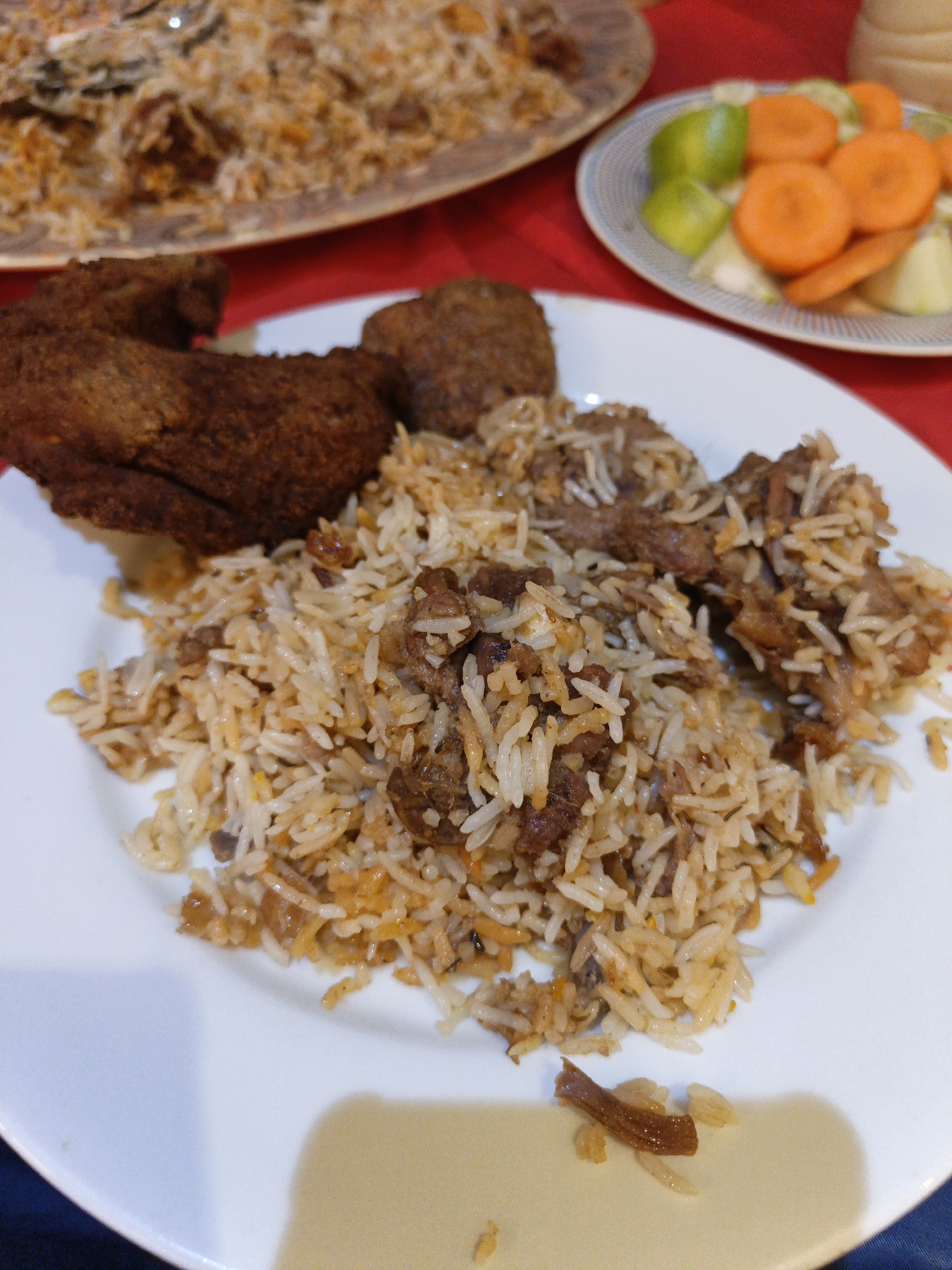 A plate of biryani, a popular rice dish in Bangladesh, is shown in close-up. The biryani, a mixture of rice, meat, and spices, is served on a white plate, with pieces of fried chicken and what appears to be a meatball or kofta on the side. In the background, another platter of biryani is partially visible, as well as a small plate of pickled carrots and limes. The setting appears to be a restaurant in Dhaka, Bangladesh, given the commonality of this dish and the red tablecloth. The lighting suggests it could be daytime, possibly indoors. There is no visible text in the image. The focus is on the food, which looks appetizing and richly prepared. The overall scene conveys a sense of casual dining and enjoyment of traditional cuisine.