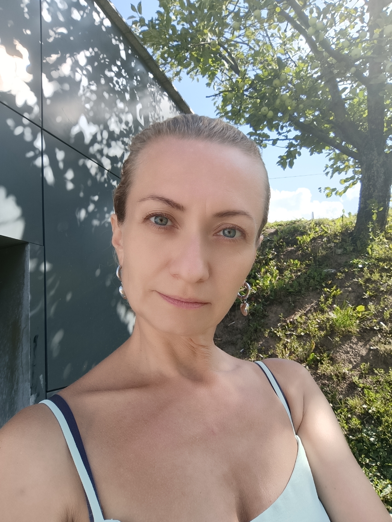 A woman is taking a selfie outdoors. She has blonde hair pulled back and is wearing a light blue top with white and navy straps. She has on hoop earrings, one of which has a heart-shaped charm. She is standing in front of a dark, textured wall, with dappled sunlight on it. To her right, there is a grassy slope with a tree bearing green fruit. The sky is blue with some white clouds. The scene appears to be a park or garden in a city, possibly Moscow, Russia given the prompt.
