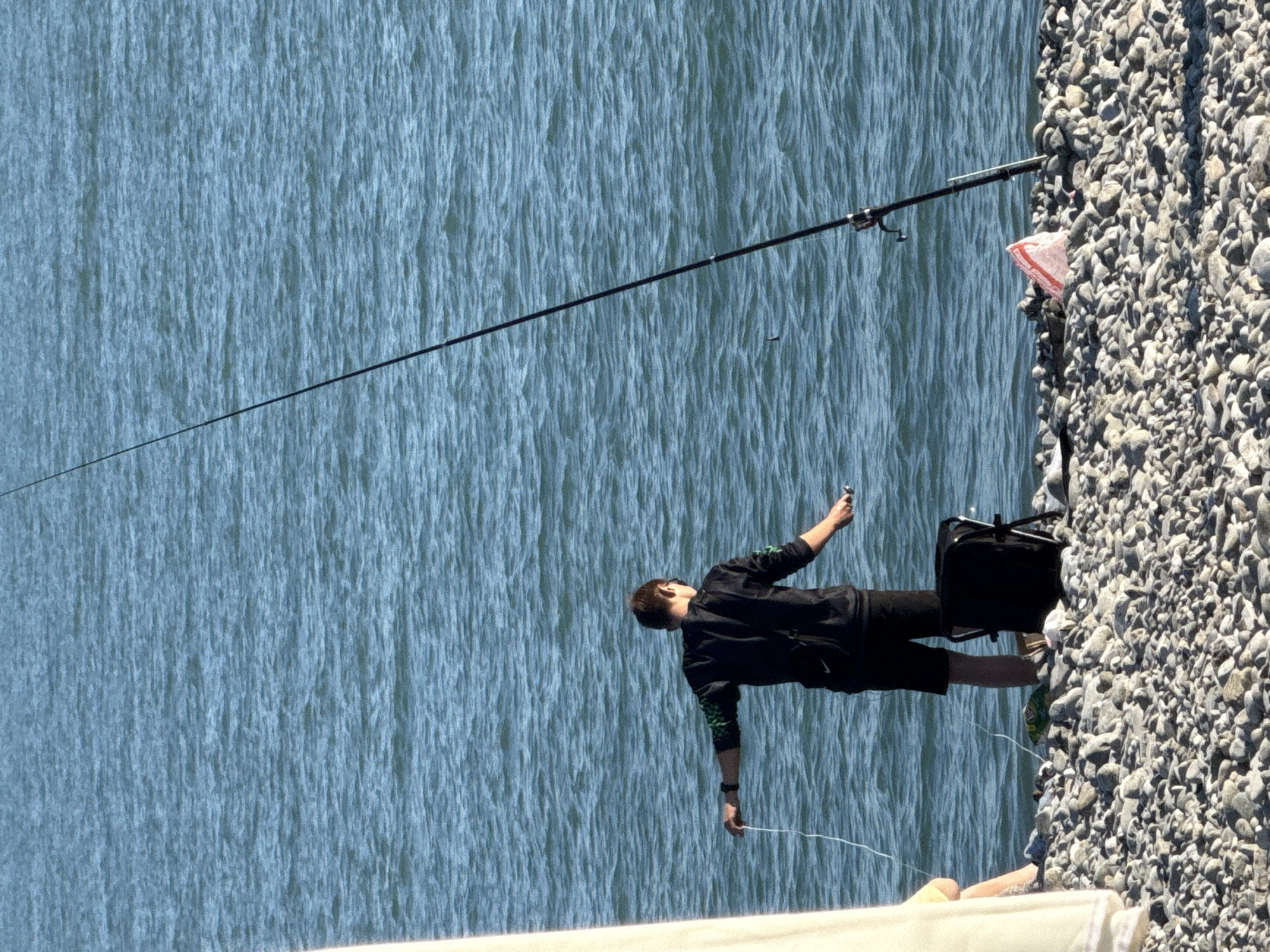 The image depicts a daytime scene on a rocky beach in the city of Sirius, Russia, where a person is engaged in fishing.
In the center, a male individual stands in shallow, calm blue water, facing away from the camera. He wears a dark long-sleeved top, possibly with green detailing on the sleeves, and dark shorts. His left arm is extended, holding a white fishing line, while his right hand holds a small, dark object, likely a component of fishing tackle.
To the right, a tall, black fishing rod is firmly planted upright among the grey and white pebbles of the beach, extending significantly upwards. A fishing reel is attached to its base. A small, dark object, possibly a lure or bobber, is visible in the water to the left of the fishing rod.
On the far left, partially obscured by a light-colored vertical structure, the lower leg and foot of a second person, likely seated on the beach, are visible.
The beach itself is entirely composed of various sized grey and white pebbles. Adjacent to the person fishing, a dark, small, foldable chair or bag rests on the pebbles. Several discarded items are present on the beach: a green and yellow snack bag lies near the chair, and a crumpled red and white plastic bag is situated further right, near the base of the fishing rod. The water exhibits a ripple pattern consistent with light waves or currents.