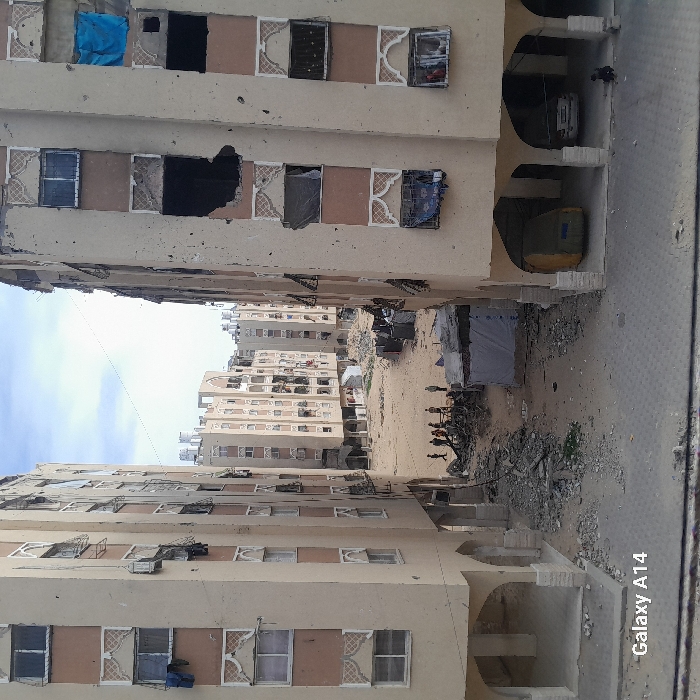 The image captures a narrow, unpaved street or alleyway flanked by multi-story residential buildings, stretching into the distance under a partly cloudy sky. The overall scene suggests significant damage and disrepair.
On both the left and right sides of the foreground and midground are tall, light-colored (beige or light brown) apartment buildings. These buildings exhibit extensive damage: numerous windows are shattered, missing, or boarded up, revealing dark interior spaces. Some window frames are broken. Decorative patterns, possibly made of concrete or plaster, adorn the areas around many windows, adding a touch of faded ornamentation to the damaged facades. On the lower levels of the building on the left, some window openings are covered with blue tarps or fabrics. Near the bottom left, a person wearing a blue shirt is visible peering out from a window. The building on the right has ground-floor archways that seem to lead to sheltered areas, under which a white car and a yellow or olive-green container or vehicle are parked.
The ground between these buildings is a rough, unpaved expanse of dirt, sand, and significant amounts of rubble, concrete fragments, and assorted debris. In the middle distance, on this unpaved ground, a group of approximately eight to ten people, many appearing to be children or young individuals, are gathered. They seem to be interacting with the environment or each other, with some standing and others possibly crouching. Adjacent to this group, a makeshift shelter or tent made of white fabric with dark supports stands on the debris-strewn ground.
Further into the background, more multi-story buildings, similar in style and condition to those in the foreground, line the alleyway, extending towards the horizon. The sky above is a pale blue with scattered white clouds, indicating it is daytime. On the far right edge of the image, a small section of what appears to be a paved road or sidewalk is visible.
In the bottom right corner of the image, the text "Galaxy A14" is visible, indicating the device used to capture the photograph. The scene conveys a sense of urban decay, potential conflict, or long-term neglect, with signs of makeshift living amidst damaged structures.