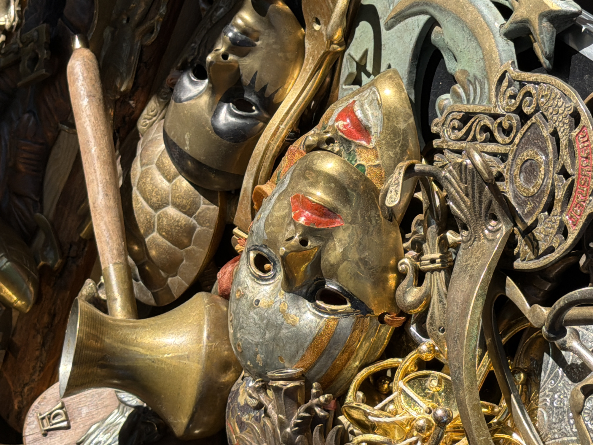 This image presents a close-up, top-down view of a dense assortment of brass and metallic decorative objects, likely displayed for sale at an outdoor market stall in Riga, Latvia. The scene is characterized by bright, direct sunlight, creating strong highlights and pronounced shadows.
In the upper-left section, a polished brass coffee pot, designed in the cezve style with a fluted texture, stands prominently. Extending diagonally from the upper-center is a rounded wooden handle connected to a brass component, suggesting a part of a larger tool or decorative implement. Below this handle, a brass ornament shaped like a segmented turtle shell is visible.
The central area features several stylized metallic masks. The most dominant mask, situated centrally, exhibits a greenish-grey patina accented with gold bands, dark eye openings, and two distinct red-painted lips. Behind it, a second golden mask with dark, stylized markings around the eyes is partially discernible. A third, predominantly golden mask with red accents is visible in the lower-right quadrant.
In the lower-center, a detailed metallic Hamsa (Hand of Fatima) pendant, incorporating an eye design and a fish motif, bears a red inscription along its lower edge. Adjacent to it, a crescent moon and star ornament, displaying a greenish patina, is present. The collection includes various other ornate metallic pieces, chains, and smaller decorative elements, all exhibiting varying degrees of tarnishing and patina, consistent with aged or antique-finished metals. The underlying surface appears to be wood, indicative of a market display. No individuals or active interactions are depicted within the frame.