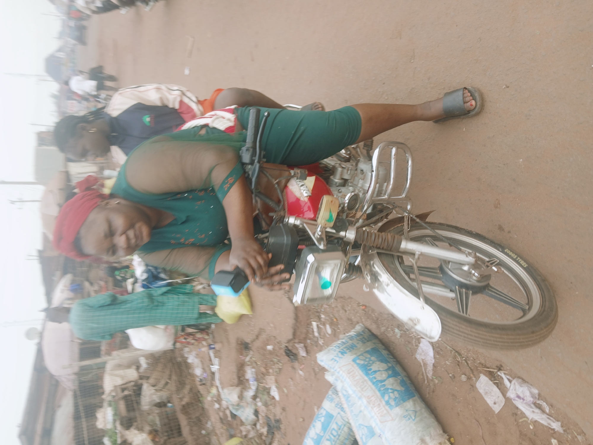 The image depicts a scene in what appears to be a market area in Ankpa, Nigeria. A woman wearing a green dress and a red headscarf is sitting on a red and silver motorcycle. Another person is seated behind her. The motorcycle is parked on what looks like a dirt or unpaved surface. Debris and trash are scattered around the base of the motorcycle, along with a large white sack printed with blue text. In the background, there are other people, vehicles, and buildings, suggesting a busy, populated area typical of a market setting.