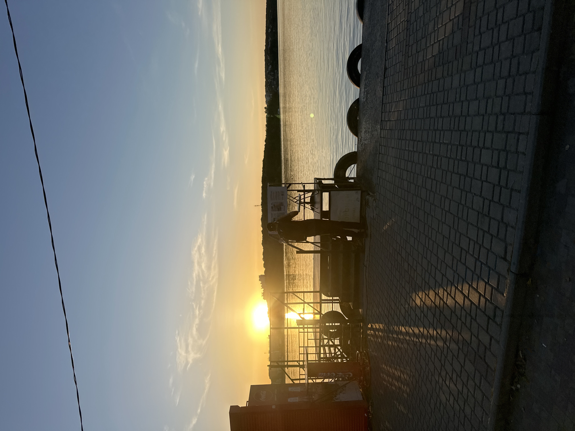 The image depicts a sunset over a body of water, possibly a river or lake, with a silhouette of a person standing on a pier. The person, wearing a cap and backpack, appears to be taking a photograph or recording a video. The pier is paved with cobblestones and equipped with metal railings and what seem to be old tires serving as fenders. A red structure, possibly a kiosk or building, is visible on the left. The sky is clear with a few wispy clouds, and the setting sun casts a warm glow over the scene. The background shows a tree-lined shore with distant buildings. The overall atmosphere is tranquil and serene. While no specific landmarks are identifiable, the scene suggests a waterfront urban or suburban setting.