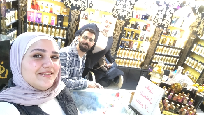 The image depicts three individuals inside a brightly lit perfume shop located in Barza Municipality, Syria. In the immediate foreground, a female with light skin, wearing a light-colored hijab and a dark vest over a white top, smiles directly at the camera, appearing to take a selfie. Behind her and slightly to the right, a male with a beard and glasses, dressed in a blue and white plaid shirt, smiles towards the camera. To his right, a second female wearing a light-colored hijab and dark clothing is partially visible, also smiling at the camera, with a light-colored handbag resting on her lap or arm. The background consists of dark-paneled display walls with ornate gold decorative elements and columns, filled with numerous perfume bottles. The lower sections of the displays show rows of uniform gold-colored bottles, while upper shelves feature various other perfume products in different containers. A glass-topped counter is present in the foreground and mid-ground, displaying a white sign with red Arabic text (reading "لتبقى مملكتنا كل ما طاب") and a multi-tiered stand holding numerous small, colorful perfume bottles.