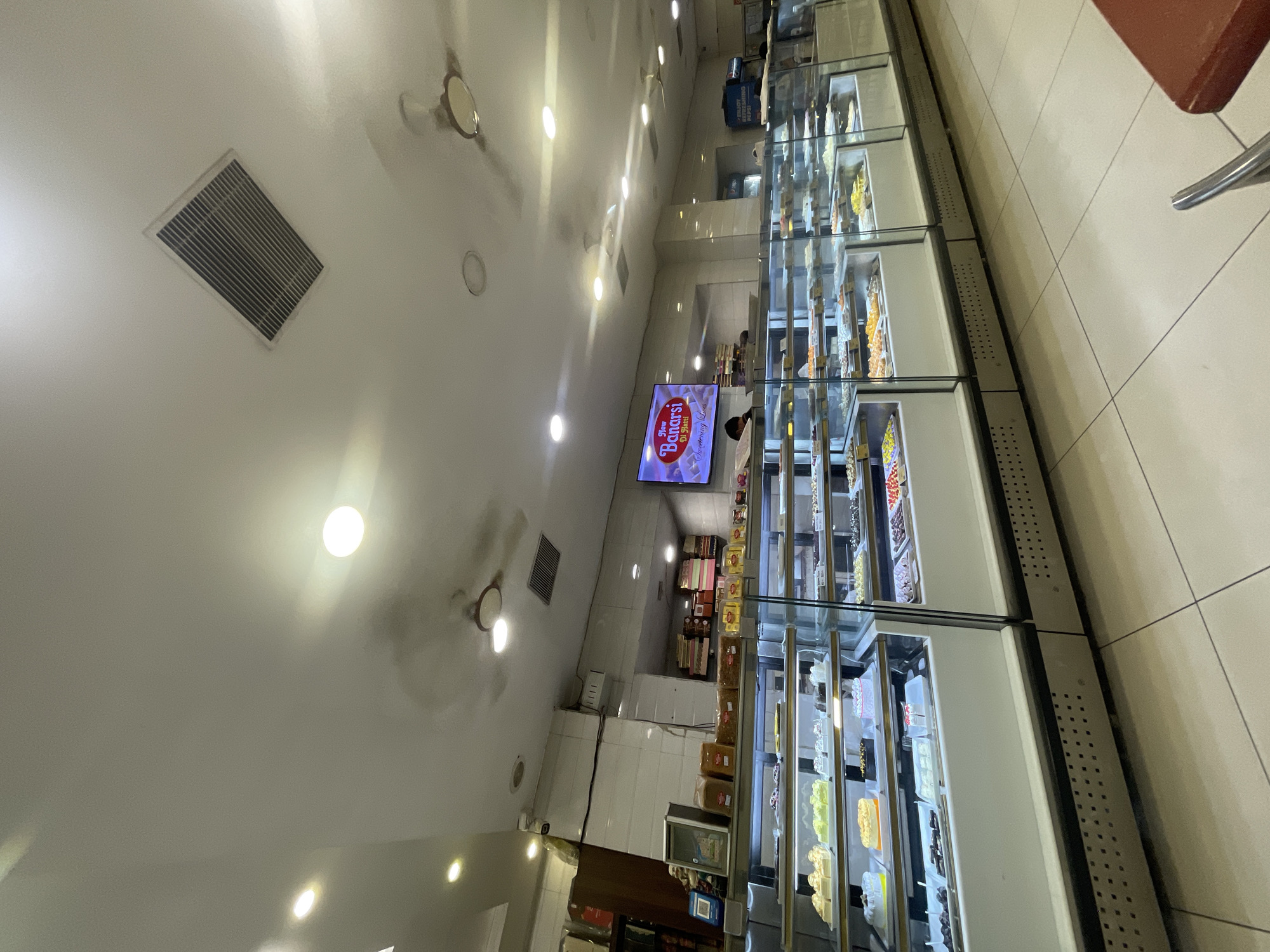 The image captures the interior of a sweets shop, likely in Pathankot, India, given the visible branding. The shop features a prominent display of various sweets and cakes arranged in refrigerated glass cases. A large screen on the wall displays an advertisement for "New Banarsi Di Hatti - Sweetening Lives." The ceiling has multiple light fixtures and two ceiling fans. In the background, there are shelves stacked with boxes and a beverage cooler. The overall environment appears clean and well-lit, designed to showcase the products.
