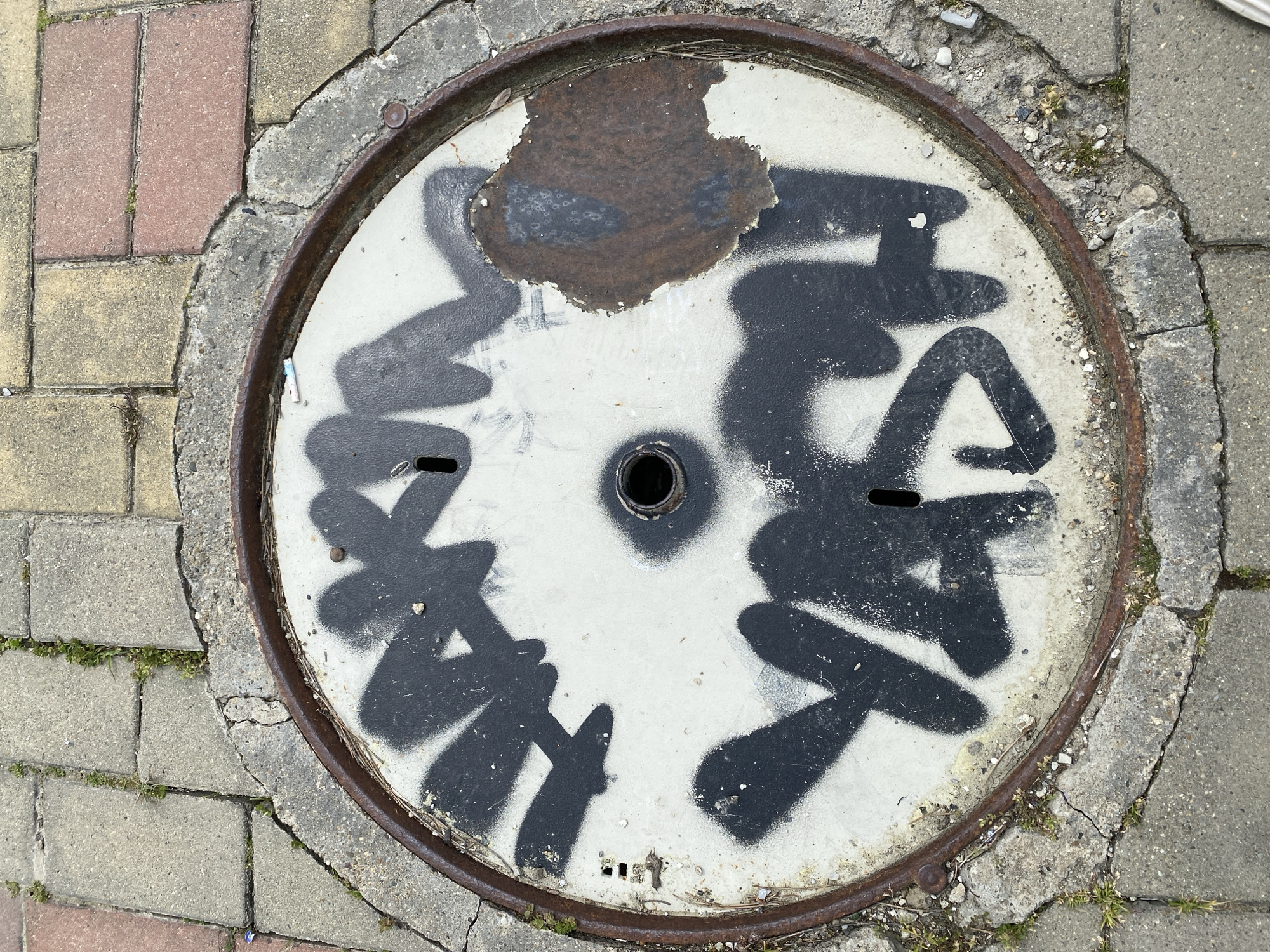 The image captures a circular manhole cover set into a paved surface in Gelendzhik, Russia. The manhole cover itself is primarily light grey or white, made of concrete or metal, and features a robust, rusted brown metallic rim. The surface of the cover is heavily adorned with black spray-painted graffiti, consisting of stylized, thick lines and shapes that appear to be illegible tags, covering much of the light-colored surface. A prominent area of dark brown rust and chipping paint is visible on the upper portion of the cover. There is a small circular hole in the very center and two elongated oval holes, likely for lifting, positioned symmetrically on either side of the center.
The surrounding area is a patchwork of interlocking pavers or bricks. Directly adjacent to the manhole's metal rim is a rough, cracked concrete border, which then transitions into the pavement. The pavers vary in color, with some appearing reddish-brown and others light grey or yellowish-grey, indicating different types of stone or wear. Weeds and small patches of grass are growing in the cracks between the pavers and the concrete border, suggesting a somewhat unkempt or aged environment. Small bits of gravel, dirt, and dried organic matter are scattered around the edges of the manhole and within the pavement cracks.
The scene depicts a static outdoor urban or suburban setting, likely during the daytime, given the bright and even lighting, though no distinct shadows are visible to indicate strong direct sunlight. There are no people present, and no specific activity is taking place other than the evidence of past human interaction (graffiti) and natural processes (wear, rust, plant growth).