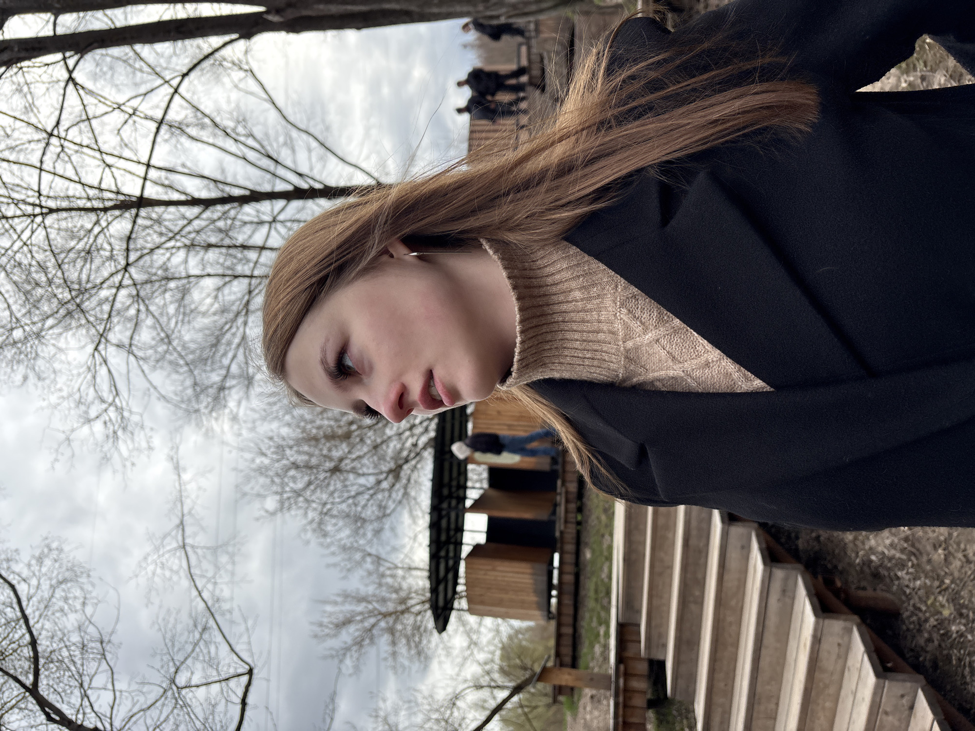 The image depicts a young woman in an outdoor setting. She is wearing a light-colored turtleneck sweater and a dark coat. The background features bare trees, a cloudy sky, a wooden structure resembling a building or platform, and a set of stairs. The overall impression is that the location is in a natural, perhaps park-like, area, and the photograph is taken at an angle, causing the scene to appear tilted. There is a group of people at the platform above. The location is in the city of Sdt Shcherbakovka, Russia.