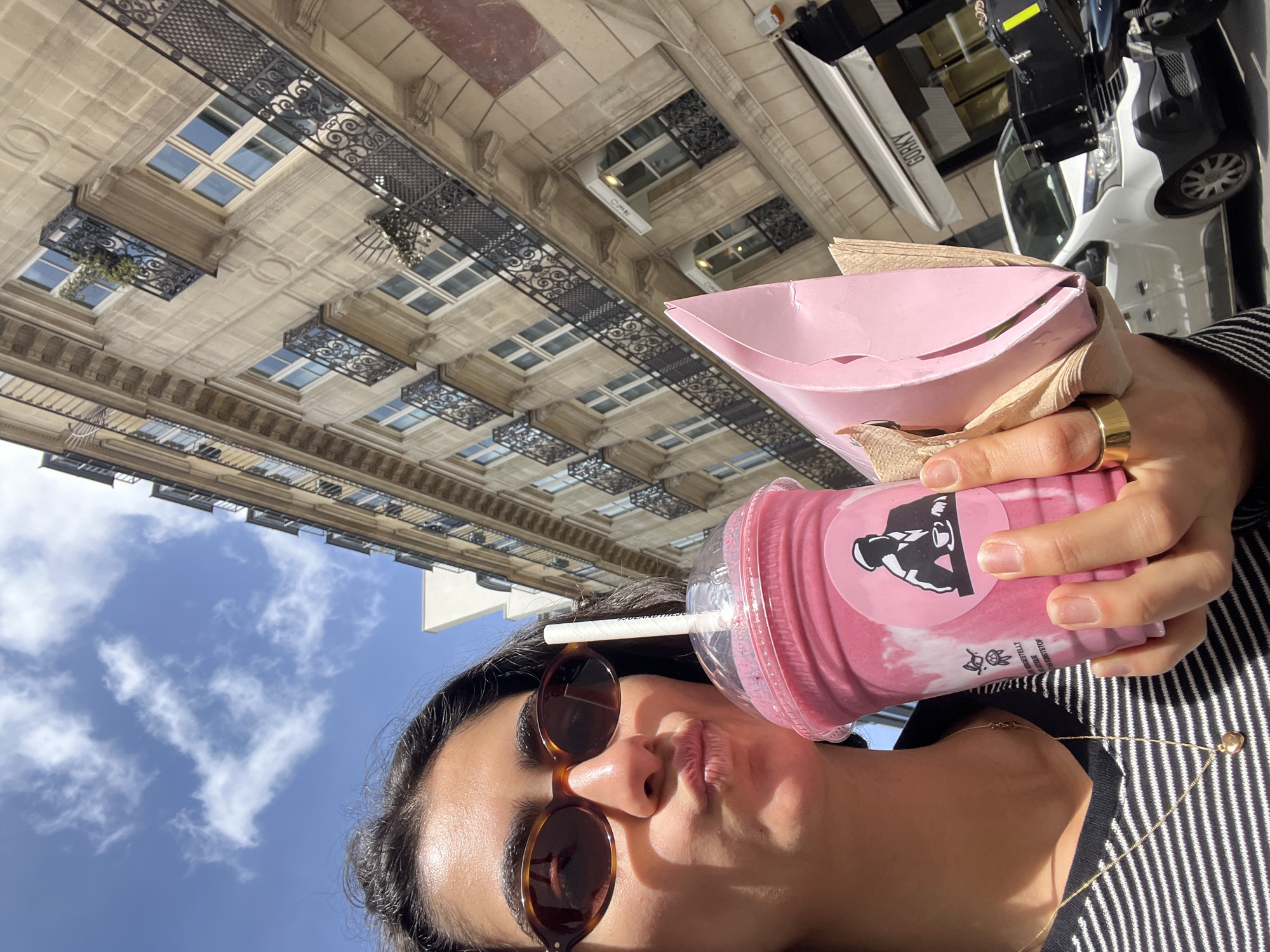 A low-angle, rotated selfie captures a person holding a pink drink and a matching container against the backdrop of a sunny Parisian street. The subject, seen from the nose up, wears dark tortoise-shell sunglasses and has their lips playfully pursed. They are wearing a black and white striped top and a delicate gold chain necklace, with a gold band ring visible on their right hand.
In their hand, they hold a clear plastic cup filled with a bright pink liquid (likely a smoothie or shake), featuring a black logo of a figure, possibly a weightlifter, and a small bunny-like emblem. A white paper straw protrudes from the cup. Beside the drink, a larger, light pink paper container, possibly for food, is held, with crumpled brown paper napkins tucked inside.
The background showcases classic Parisian architecture: a grand, light-colored stone building with multiple floors, each adorned with ornate black wrought-iron balconies and window railings. The sky above is a clear, vibrant blue with scattered white clouds, indicating a bright day. In the upper right corner, parts of a street scene are visible, including a white van, a black vehicle, and a white commercial sign that clearly reads "GORKY". The image conveys a casual, enjoyable moment in Paris.